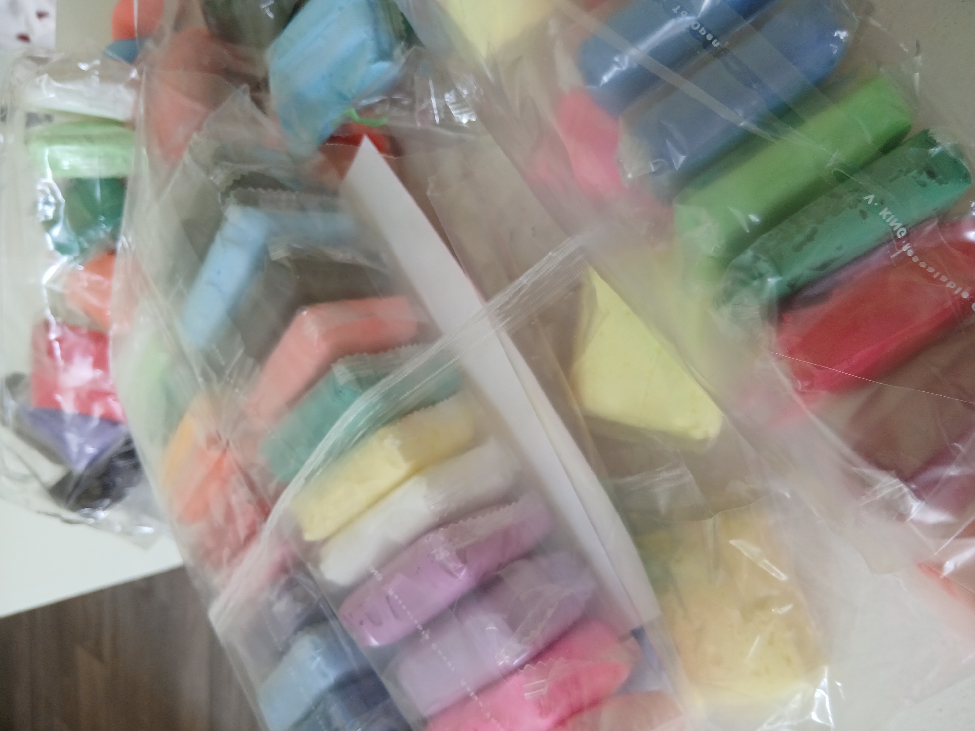 The image displays a close-up view of numerous colorful blocks of what appears to be modeling clay or dough. The blocks are individually wrapped in clear plastic and then grouped together inside larger transparent plastic bags, possibly for storage or sale.
A wide array of colors is present, including various shades of blue, green, red, yellow, white, pink, purple, black, and orange. In the central and lower portions of the image, rows of pastel-colored blocks (pink, purple, white, yellow, light blue) are visible within one of the larger bags. Another large bag in the upper right contains brighter hues like dark blue, light green, dark green, and red.
The setting appears to be indoors, possibly on a light-colored table or surface. The lighting suggests an artificial indoor environment, with reflections visible on the plastic packaging. The image is somewhat blurry, particularly towards the edges, but the general forms and colors of the blocks are distinct.
No specific activity is taking place; the image is a static shot of the packaged materials. No people are visible.
Visible text, though inverted and partially obscured by reflections and blur, can be seen on some of the individual wrappers. On a green block wrapper in the upper right, the words "A. KING" and "MODELING CLAY" are discernable in inverted orientation. Similar inverted text, likely also "MODELING CLAY", is visible on a blue block wrapper in the upper central area.