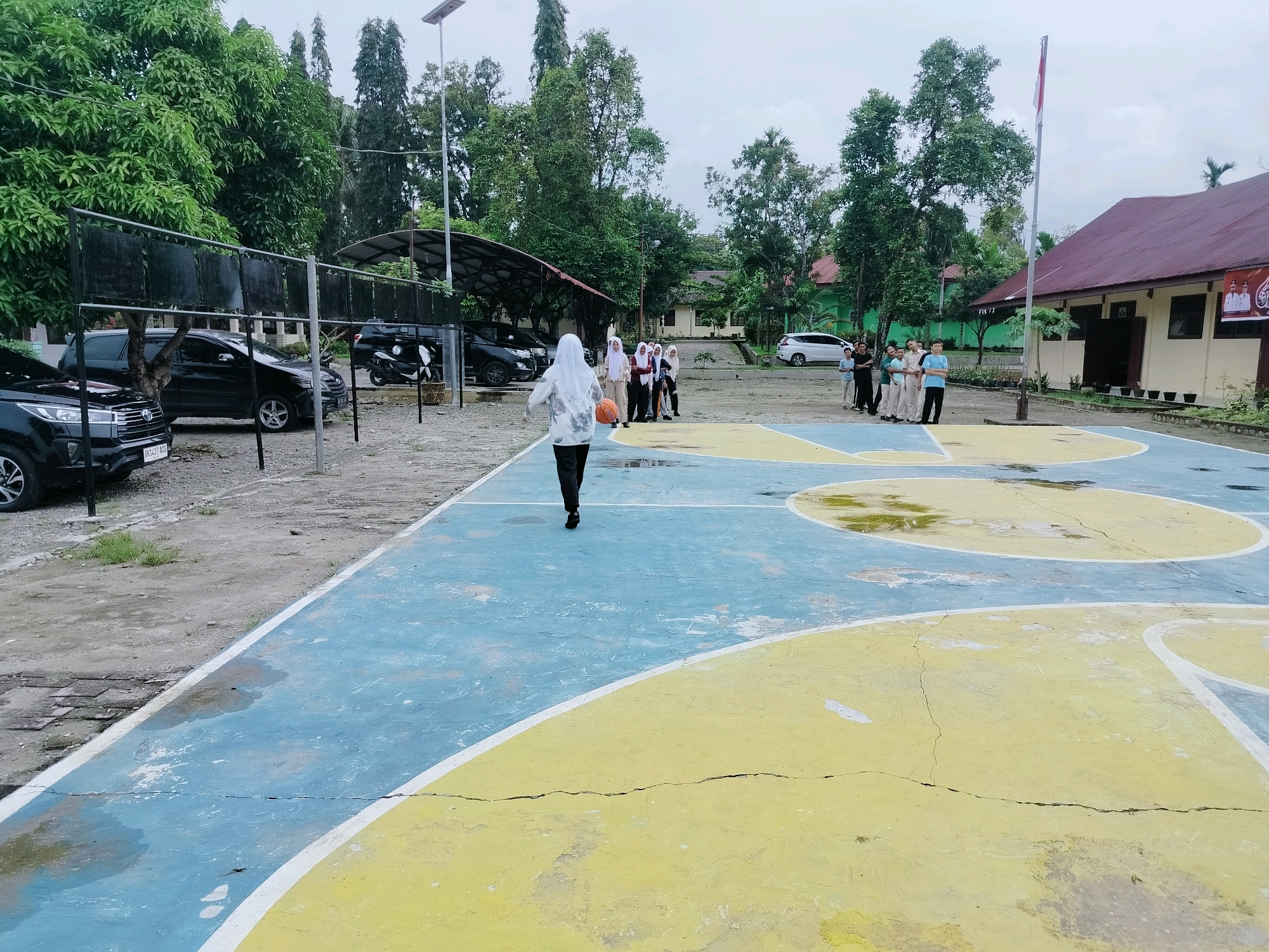 In Lawebekung, Indonesia, a group of students are on a basketball court, which is wet and cracked, with yellow and blue painted sections. A female student in a hijab is dribbling a basketball, facing away from the camera, while several other students stand in a huddle to her right. In the background, cars are parked under a canopy to the left, and a building with a red roof stands to the right, adorned with banners. Trees and a cloudy sky complete the scene.