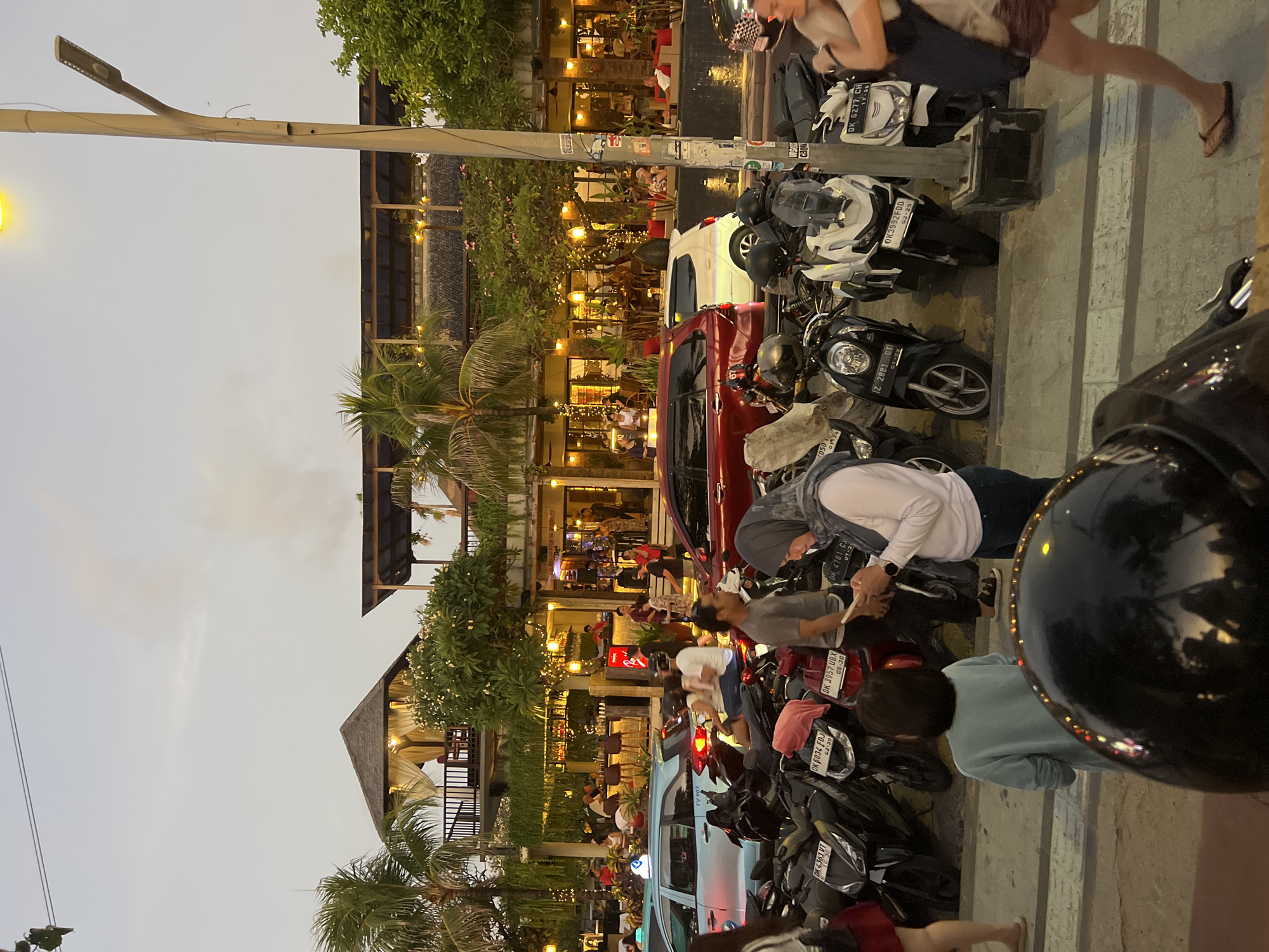 This image captures a bustling street scene in Kuta, Indonesia, likely during the late afternoon or early evening. The foreground is dominated by a parked row of motorcycles, some with helmets resting on them. A black helmet, reflecting the ambient light and the twilight sky, is prominently positioned in the lower part of the frame.
In the middle ground, several people are visible. A woman wearing a hijab and a white shirt is looking at her phone. To her left, a person in a light blue long-sleeved shirt stands facing away from the camera, while another individual in a grey t-shirt is also present. Further back, a few more individuals are seen near parked cars, including a red SUV and a light blue car.
The background features a lively street with restaurants and shops, characterized by their warm lighting and outdoor seating areas. Palm trees are scattered throughout the scene, adding to the tropical atmosphere. A street lamp with a lit bulb stands tall on the right side of the image, illuminating the area. The sky is overcast with hints of color from the setting sun. The overall impression is one of a vibrant and active urban environment in Kuta, Indonesia.