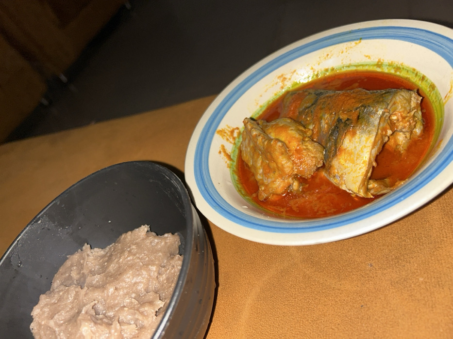 The scene, located in Egbe, Nigeria, displays two bowls of food positioned on an orange-brown, textured surface. In the upper left quadrant, a black, deep bowl contains a substantial amount of light brown, textured, semi-solid food. To the lower right, a white ceramic bowl with blue and green concentric stripes on its rim holds several pieces of fish, which are largely submerged in a vibrant reddish-orange stew or sauce. A distinct diagonal shadow line traverses the background, suggesting a directional light source illuminating the left side of the frame and casting the upper right area into darkness. No individuals are present in the image.