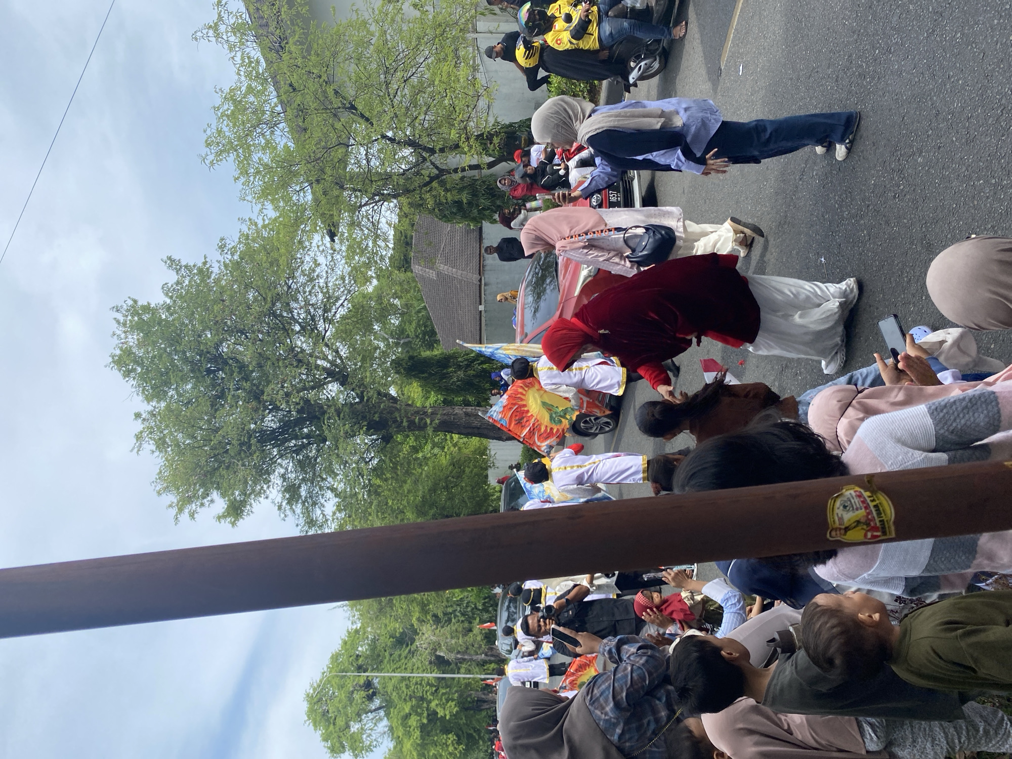 The image depicts a street scene in Banda Aceh, Indonesia, possibly during a cultural event or parade. In the foreground, a large wooden pole, possibly a street signpost, is prominently featured and slightly obscures the view. A crowd of people, including children and adults, are gathered on the street, many facing towards the right of the frame, suggesting they are watching an event unfold. Several individuals are holding up mobile phones, indicating they are taking pictures or videos.
Further into the scene, a red car is visible, and a procession of people in traditional white and gold attire are walking alongside it. Some of these individuals are holding colorful banners or flags with intricate designs. Other people in the procession are wearing hijabs and other contemporary clothing. A person in a red velvet robe is walking in front of a group, and another individual is operating a camera on a tripod. Motorcycles and other vehicles are also present on the street. The environment includes lush green trees lining the street and buildings in the background, consistent with an urban setting in Banda Aceh. The sky is overcast with some visible clouds.