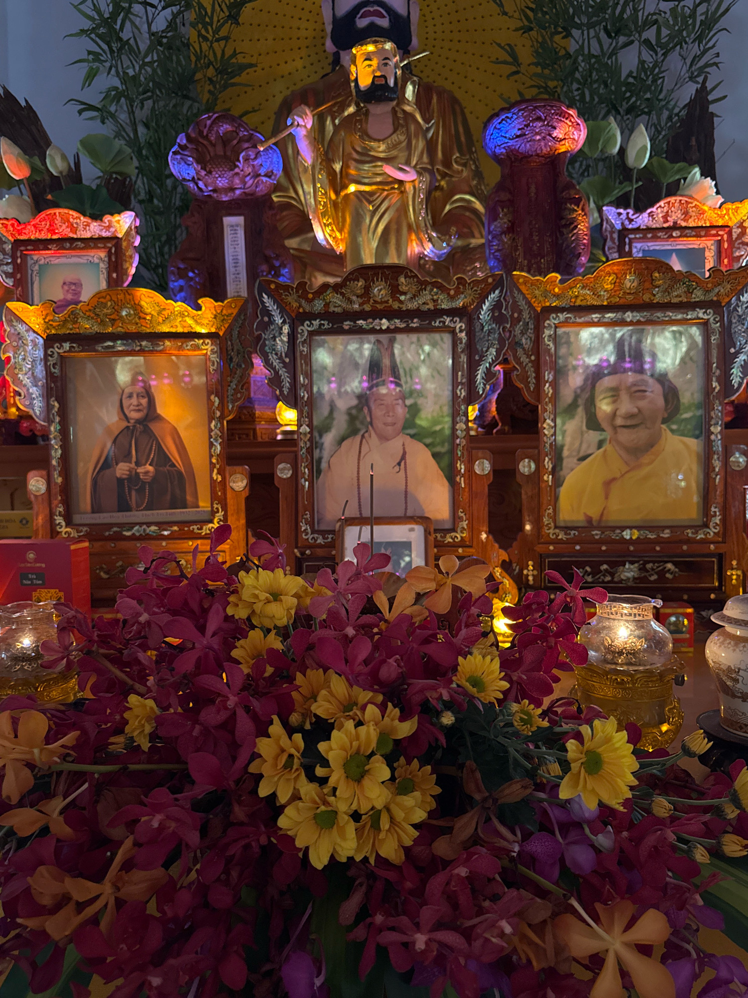 This is a medium shot, eye-level photograph taken in a dimly lit indoor setting, likely a shrine or altar. The scene is dominated by a central golden statue of a deity, possibly a Buddhist figure, with a serene expression and wearing ornate attire. Flanking the statue are three framed photographs, each depicting an individual. The leftmost photo shows an elderly person with a gentle smile, dressed in monastic robes. The central photo features a person in traditional religious attire, also with a benevolent look. The rightmost photo displays another elderly person, smiling warmly and wearing a yellow robe.
In the foreground, a lush arrangement of vibrant flowers, primarily deep red orchids and yellow daisies, creates a colorful and fragrant display. Several lit candles and ornate lanterns are placed around the altar, casting a warm glow on the scene. In the background, there are green bamboo stalks and hints of decorative elements, suggesting a spiritual or commemorative space. The overall atmosphere is one of reverence and solemnity, with a touch of warmth provided by the flowers and candles. The specific location in Nha Trang, Vietnam, is not discernible from the image itself, but the presence of Buddhist iconography and floral arrangements is consistent with religious practices in the region.