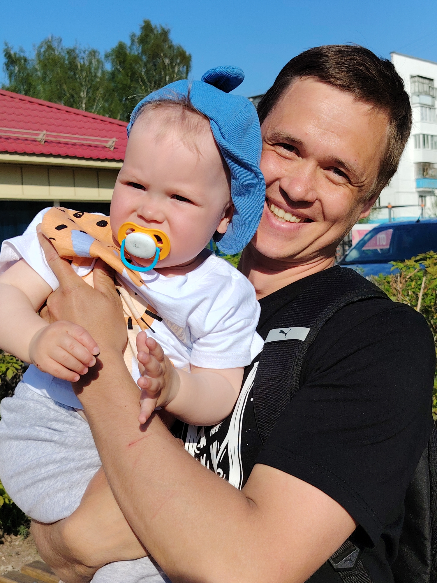 The image depicts a man holding a baby outdoors. The man has short brown hair and is wearing a black T-shirt and a backpack. He is smiling and looking directly at the camera. The baby is wearing a white T-shirt, grey pants, and a blue hat. The baby has a pacifier in their mouth and is looking at the camera. In the background, there is a building with a red roof, trees, and a blue car. The location appears to be in the city of Chaikovsky, Russia, judging by a partial registration number visible on the car (170). There are bushes and a paved area in the foreground.