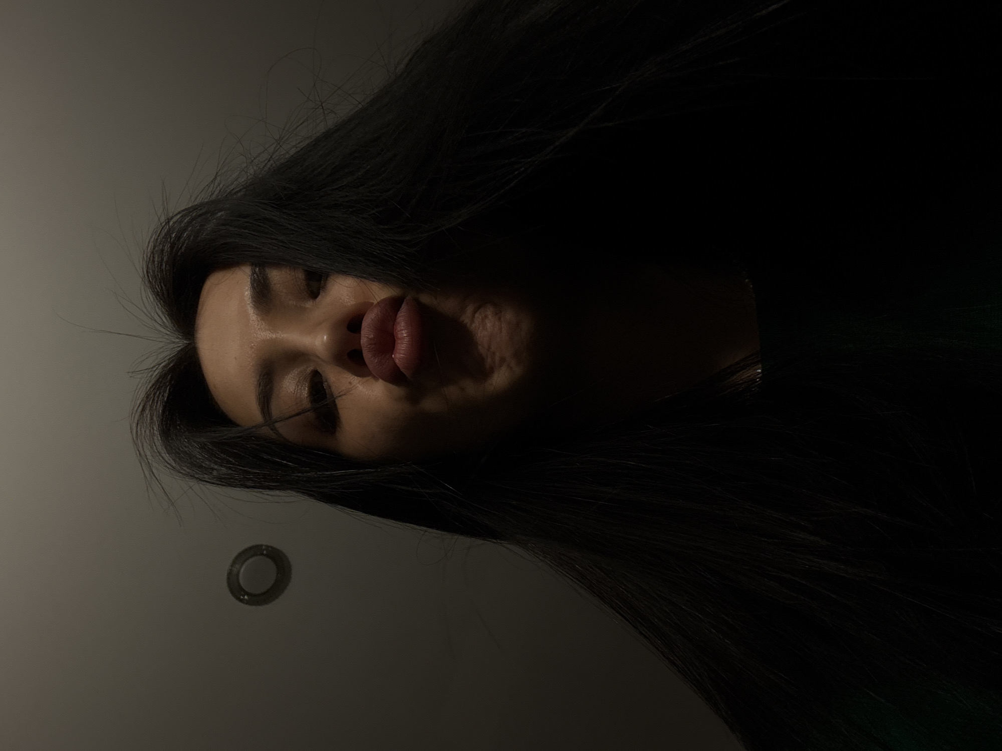 A head-and-shoulders shot of a person with long, dark hair making a "duck face." The person is angled downwards towards the camera. The background is a dimly lit white wall. A small, round object, possibly a light fixture, is visible on the wall. The location is indeterminate; no elements suggest Votkinsk, Russia.