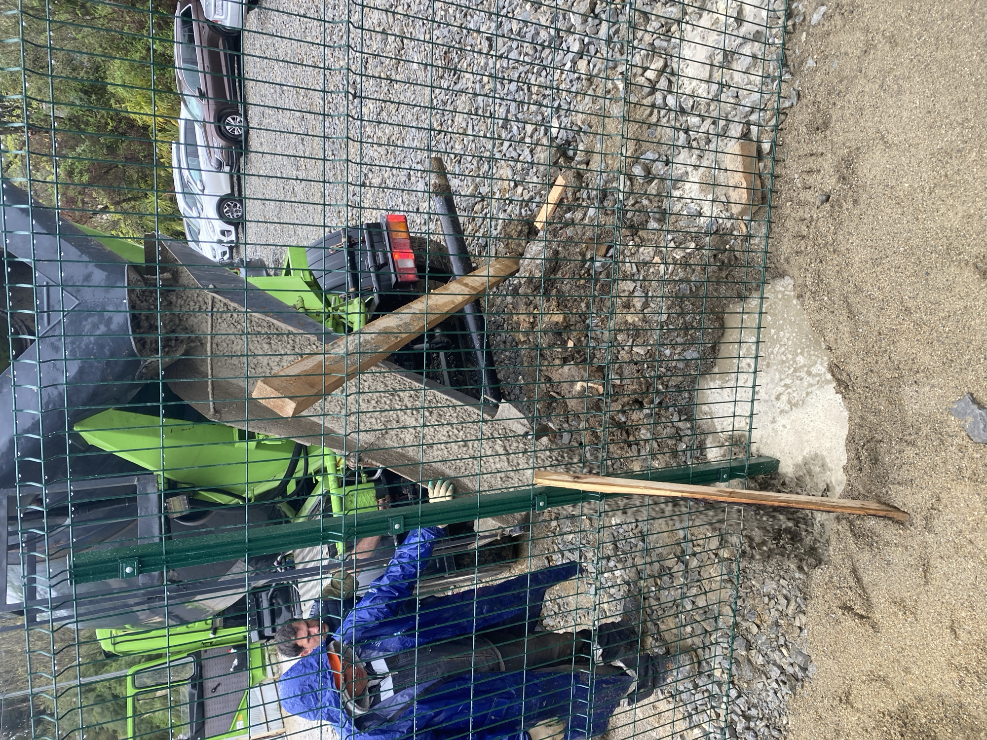 The image, likely taken in Sochi, Russia, shows a construction site viewed through a green wire fence. A man wearing a blue raincoat is present, partially obscured. A green construction vehicle with a black bucket attachment is visible behind the fence. A light-colored sedan and a dark-colored SUV are parked in the background. The ground on one side of the fence is covered in small rocks and dirt, while the other side is sandy. Two wooden planks are present, resting on the ground and partially passing through the fence. There is water pooled on the rocky side of the fence.