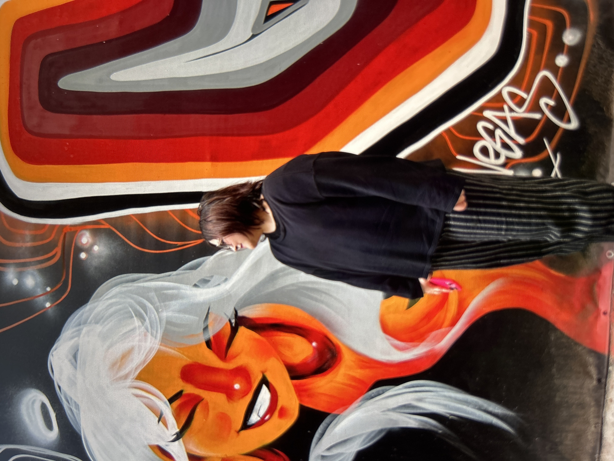 This media file features a person standing in front of a vibrant mural. The mural depicts a stylized, graphic design with concentric, wavy stripes in shades of orange, red, brown, and gray at the top. Below this, the mural transitions into an illustration of a face with white hair, orange skin, and red accents, looking upwards with a wide smile. The background of the lower portion of the mural is black with some white and orange spray-painted details.
The subject is a person with dark hair, wearing a black long-sleeved shirt and striped pants. They are positioned in the middle of the frame, leaning slightly forward and looking down towards the mural. Their right hand is extended, holding a small, bright pink object. The person's body is partially obscured by the mural's design.
There is visible text in white spray paint on the right side of the mural, which appears to be a tag or signature. The overall impression is one of urban art and personal expression, possibly an interaction with street art. The lighting suggests it is daytime, but no specific weather or time of day cues are evident beyond that. The location is not identifiable from the image.