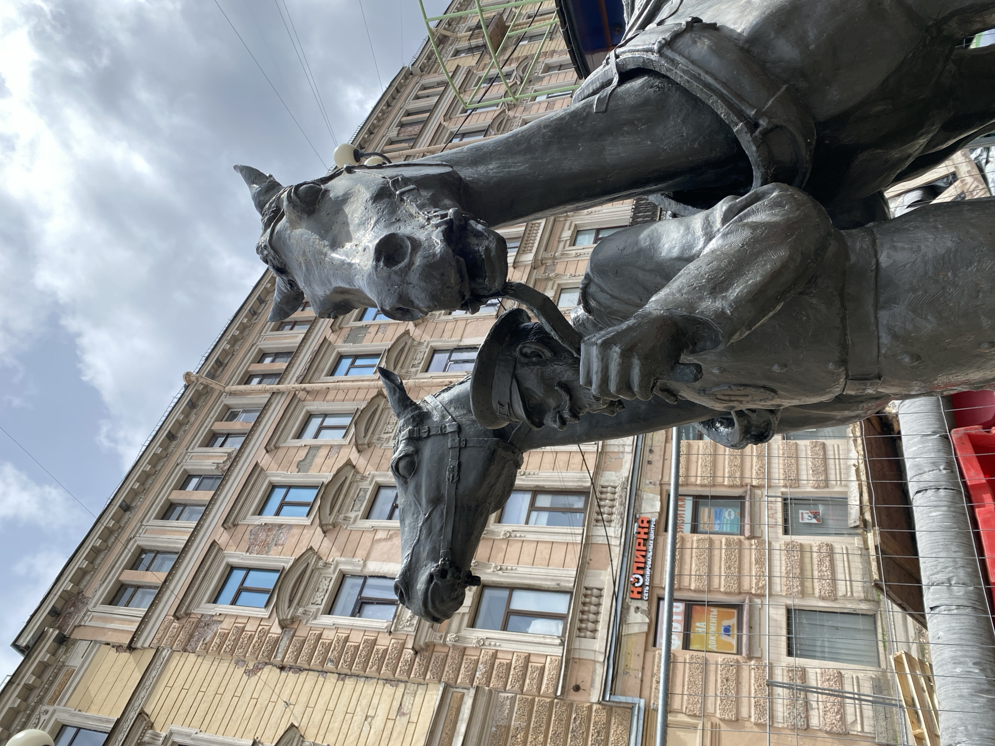 The image shows a low-angle view of the Klodt Horses sculpture on the Anichkov Bridge in Saint Petersburg, Russia. The sculpture features two bronze horse statues with their handlers. The architecture of a multi-story building is visible in the background, with repetitive windows and ornate details. Construction fencing and some scaffolding are partially visible on the right side of the frame. A sign with Cyrillic text is visible attached to the building. The sky is cloudy.