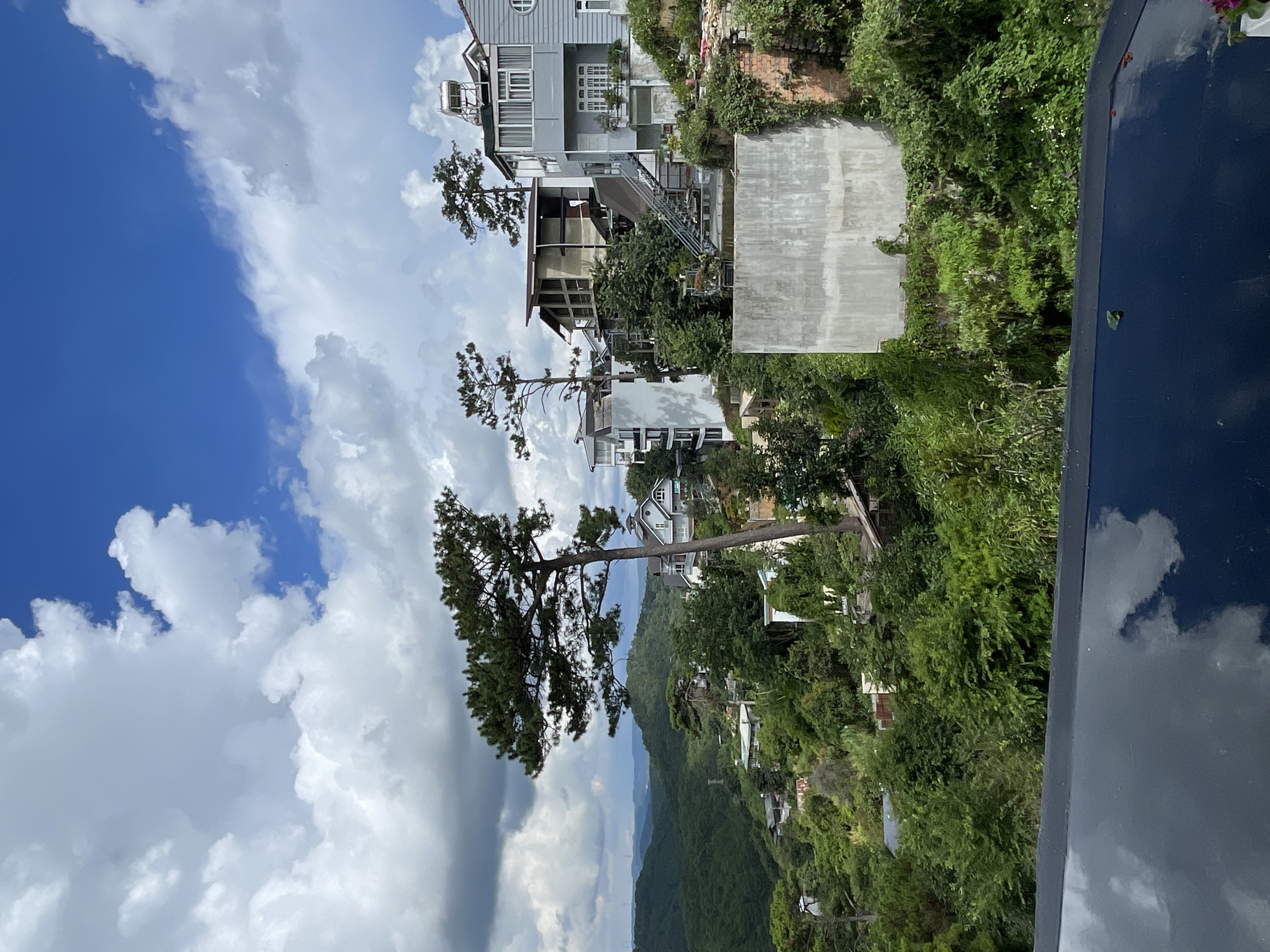 The image depicts a lush, verdant hillside in Da Lat, Vietnam, characterized by a vibrant landscape of rolling green hills covered in dense vegetation and scattered houses. The sky is a striking blue, dotted with large, fluffy white clouds that cast shadows on the landscape. In the foreground, a dark, reflective surface, likely the edge of a pool or a similar structure, occupies the bottom portion of the frame, mirroring the sky and clouds above.
The hillside is a tapestry of trees, bushes, and flowers, with a prominent, slender pine tree leaning towards the right in the mid-ground. Several multi-story houses are visible nestled into the hillside, constructed in a variety of architectural styles. Some feature white or light-colored facades with clean lines, while others exhibit more rustic brickwork and natural materials. Staircases and pathways wind up the slopes, connecting the buildings and offering access through the dense foliage. One house on the right side of the frame is a pale grey, with multiple windows and a solar water heater on its roof.
The overall impression is one of a serene, somewhat secluded residential area integrated with nature, showcasing the characteristic topography and vegetation of Da Lat. There are no people visible in the image, and the scene appears tranquil and uninhabited in the moment captured. The lighting suggests it is daytime, possibly mid-afternoon, given the interplay of sunlight and shadow.