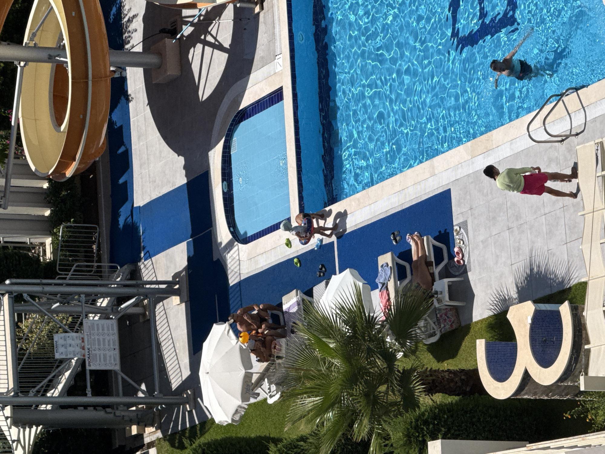 The image shows a resort swimming pool area on a sunny day in Alanya, Turkey. In the foreground, a palm tree shades a portion of a tiled patio with a unique "E" shaped water feature. Several lounge chairs are present, with some occupied by sunbathers. A white umbrella provides shade for a group of people sitting on chairs. Further back, a large blue swimming pool is visible, with two people swimming in it. Adjacent to the pool is a smaller, rectangular pool. The background features a water slide structure and lush green vegetation. The overall scene depicts a typical recreational area at a tourist destination.