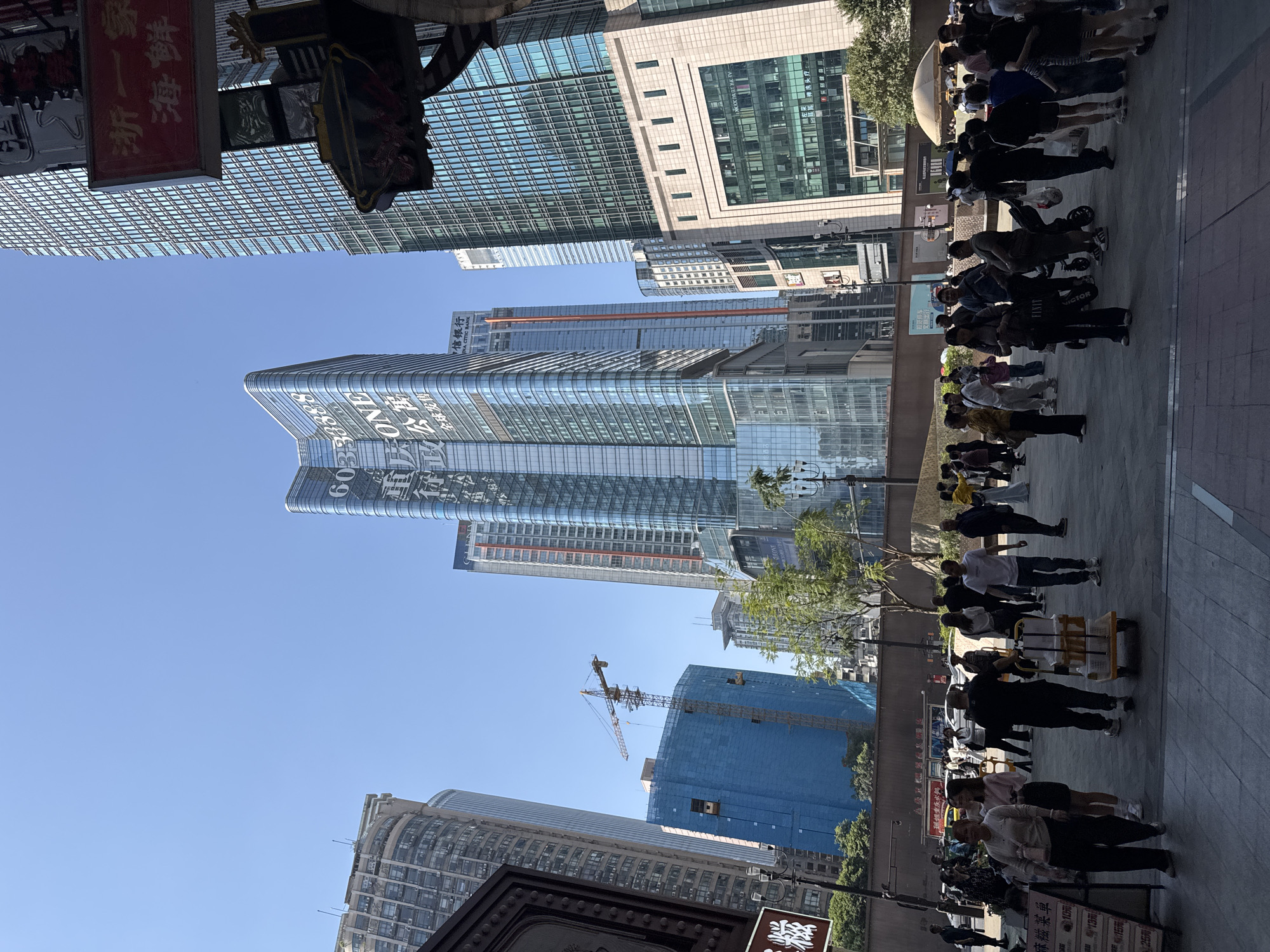 The image is a street-level shot in Yuzhong District, China. Skyscrapers rise high into a clear blue sky, including one with "ONE" and Chinese characters on its facade and another under construction, covered in blue material with a crane visible. A row of people walks along the sidewalk, appearing as a crowd. There are signs with Chinese characters in the foreground. The architecture includes traditional elements mixed with modern glass structures. The scene is daytime, with sunlight reflecting off the buildings.