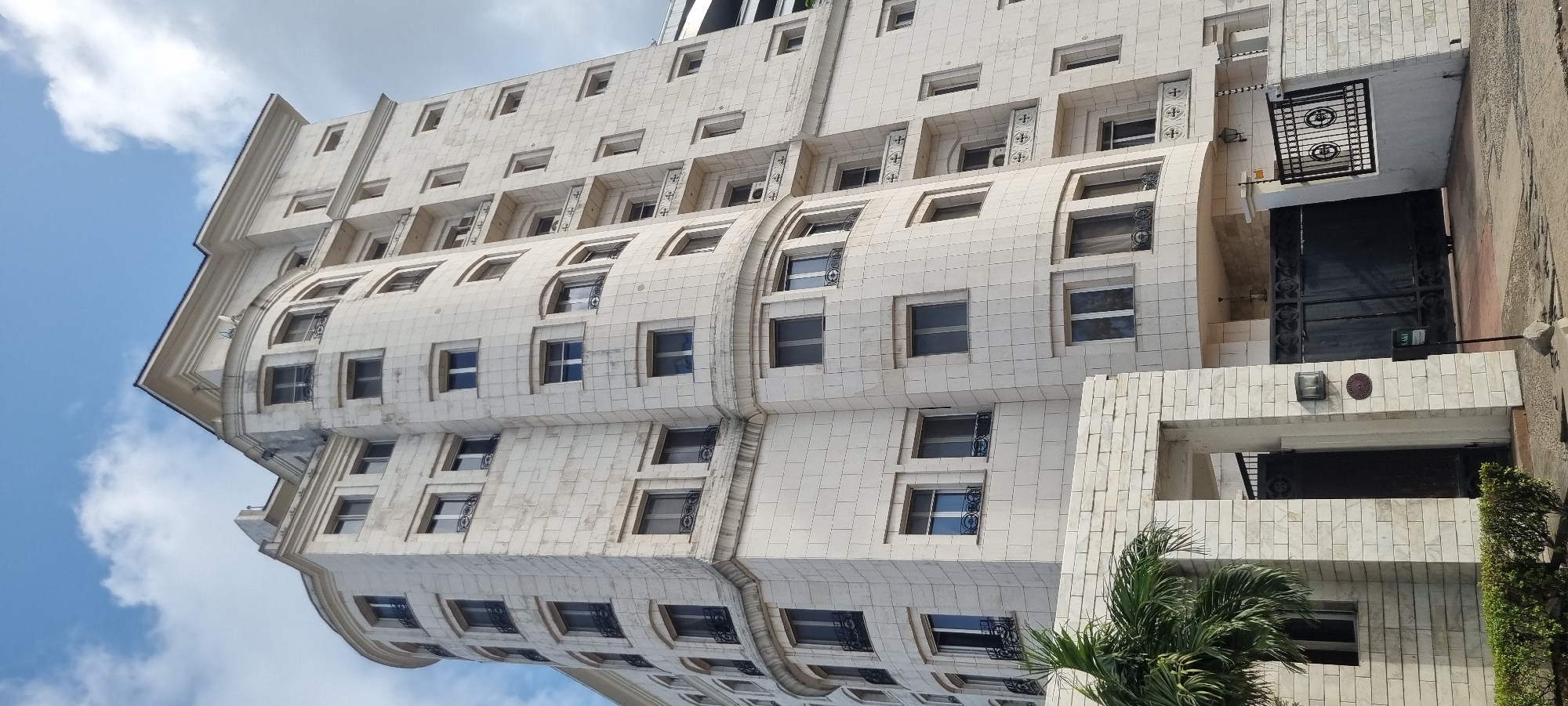 The image shows a multi-story building in Falomo, Nigeria. The building's facade is characterized by a light-colored, possibly marble-like, material. It features a mix of rectangular and curved architectural elements. Windows are consistently rectangular, with decorative ironwork outside the windows. At the base of the building, near the apparent entrance, there's a black gate and a stone structure with some greenery. The sky is visible in the upper-left, showing a blue sky with clouds. There are no people visible.