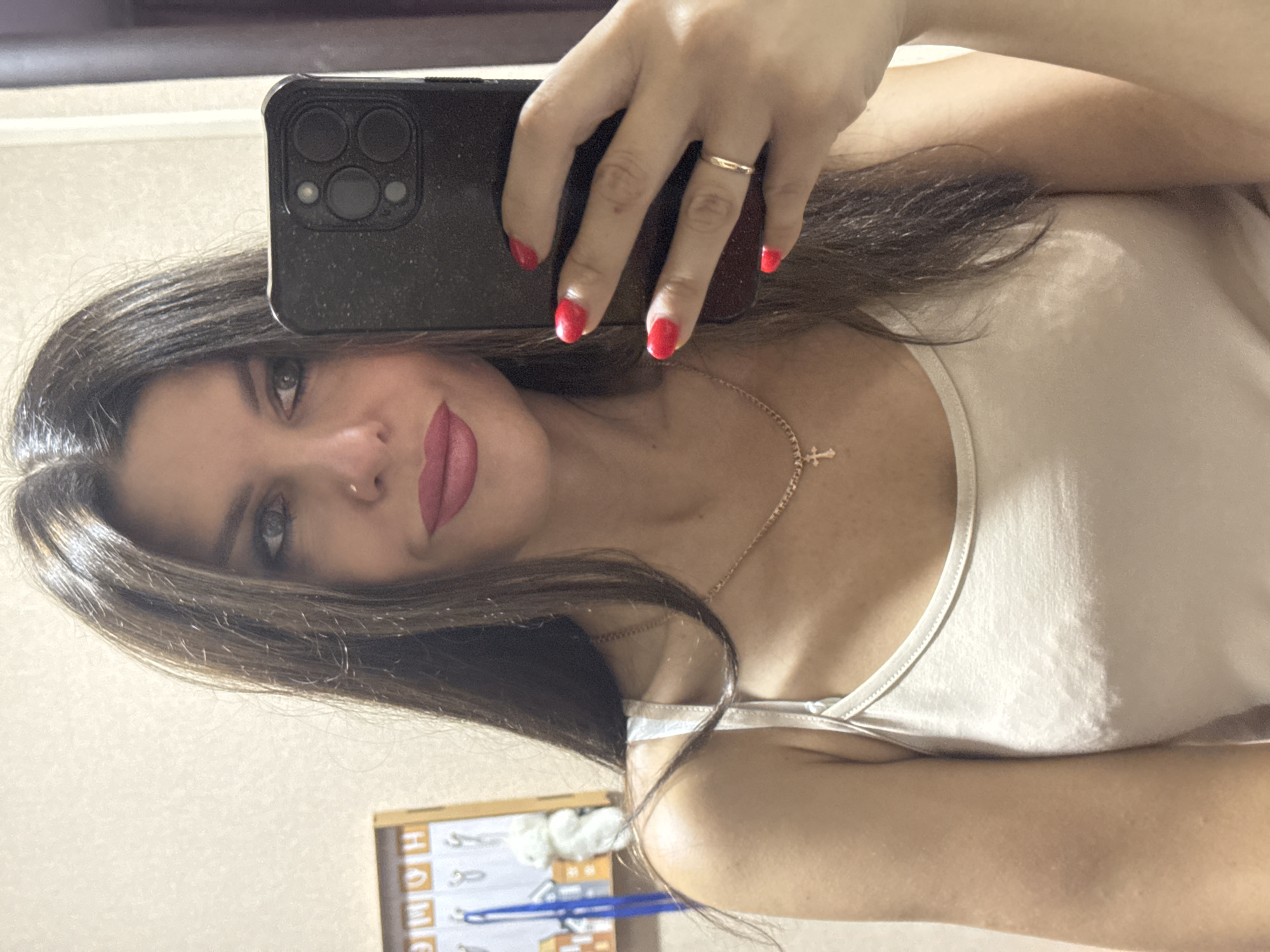 A fair-skinned woman with dark hair, red nail polish, and a white tank top is taking a selfie. She is wearing a gold necklace with a cross pendant and a gold ring on her right hand. She is holding a black smartphone in her right hand, angled to capture her reflection. In the background, a wooden frame with the word "HOME" is visible, along with some decorative items. The wall appears to be a neutral beige color. The image does not provide enough information to determine a specific location within Chaikovsky, Russia.