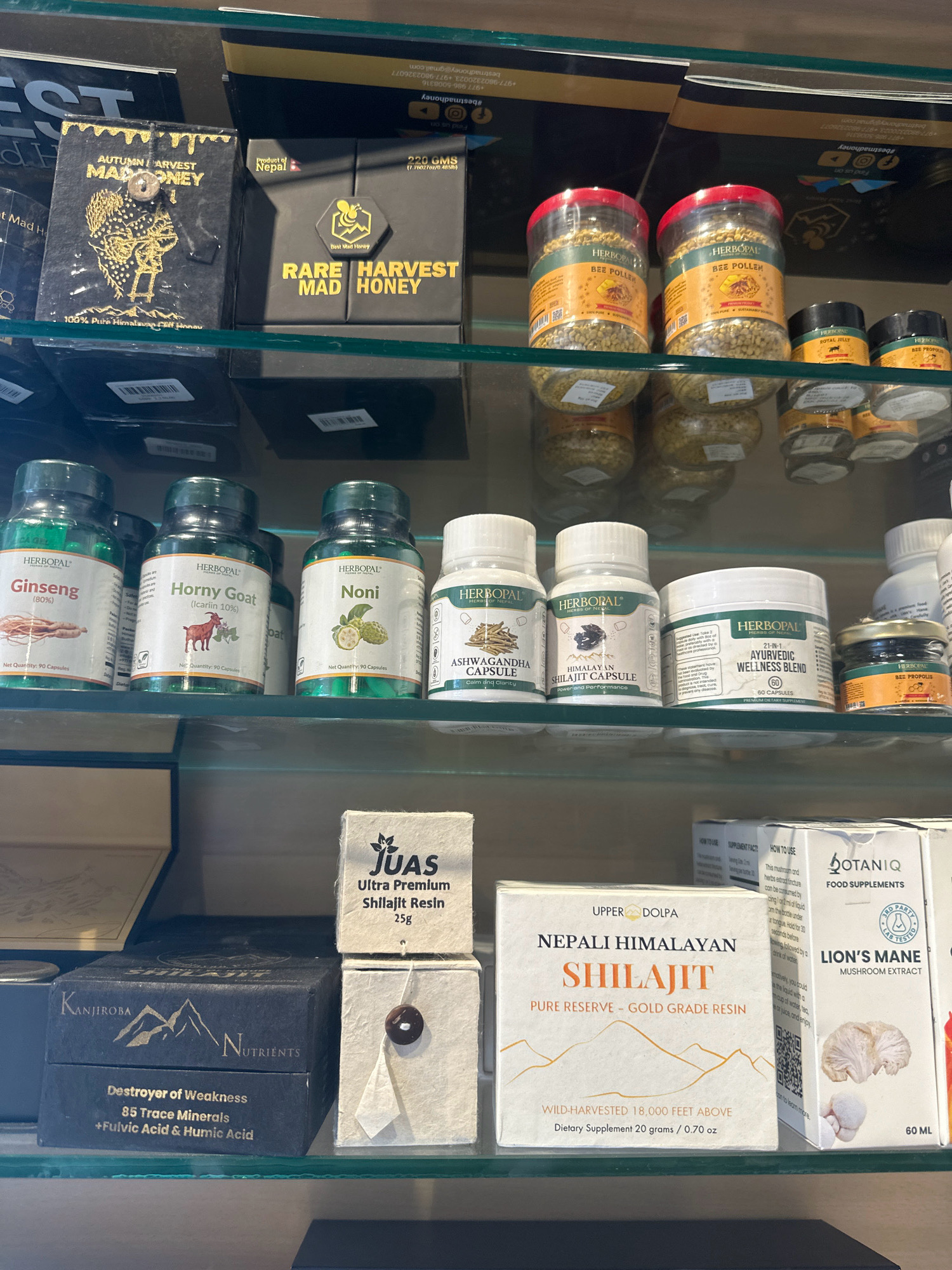 The image shows a retail display of health and wellness products on glass shelves in a store. The products include various types of honey, bee pollen, bee propolis, supplements such as Ginseng, Horny Goat, Noni, Ashwagandha Capsule, Himalayan Shilajit Capsule, and a general Ayurvedic Wellness Blend. There are also boxes of Nepali Himalayan Shilajit, and BotanIQ Lion's Mane Mushroom Extract. The store appears to be located in Lalitpur, Nepal, as indicated by product labeling such as "Product of Nepal" and brands associated with the region. The lighting suggests an indoor environment, likely a shop. No people are visible in the image.