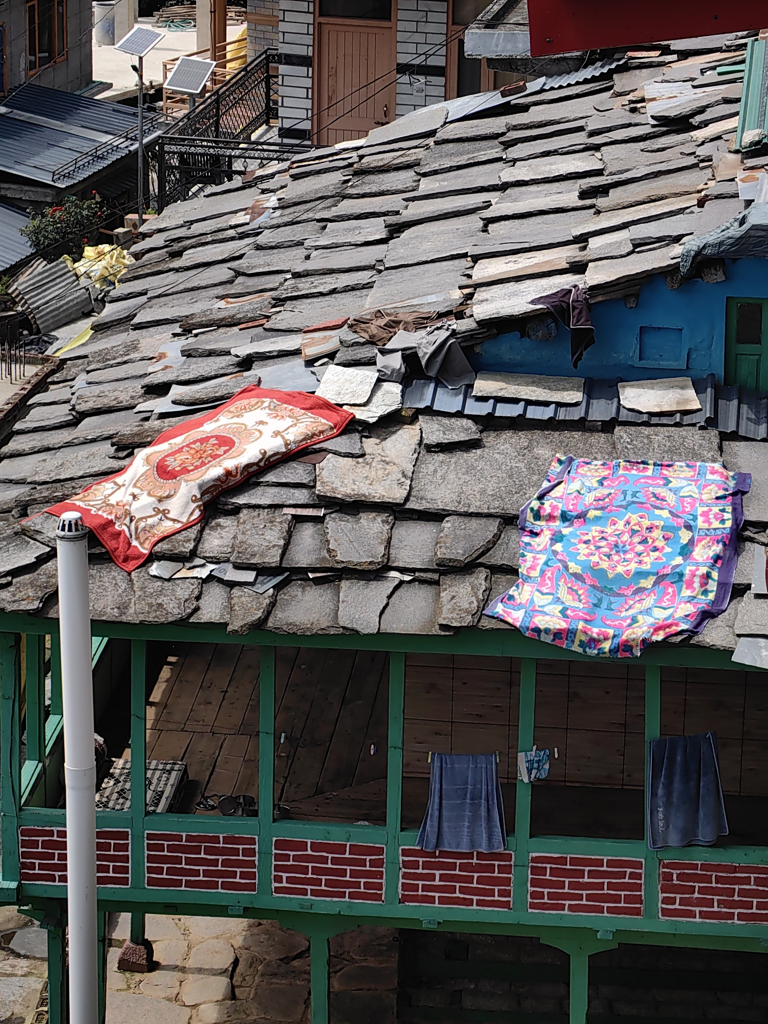 In Vashisht, India, a weathered stone-tiled roof, typical of the region, is adorned with a vibrantly patterned red and white textile and a brightly colored blue, pink, and yellow textile, suggesting they are drying in the sun. Below the roofline, a green-painted wooden balcony or porch features a red brick facade with white mortar lines. Hanging from a clothesline on this porch are several dark blue garments, one of which appears to be a towel with text. The scene is set against a backdrop of stone buildings and other rooftops, indicative of a dense residential area.