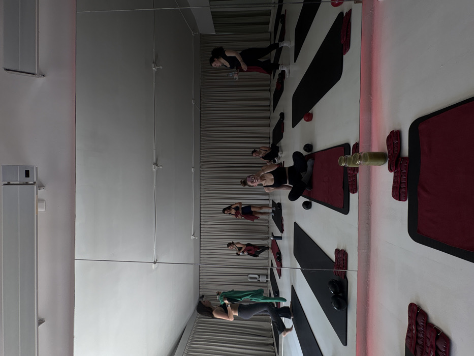 A mirrored view of a fitness studio. Several individuals are exercising on mats, some with weights or resistance bands. The mats are arranged in a line, alternating between black and maroon. Walls are mostly white. Several bottles are placed next to one of the mats. Vertical metal bars divide the wall on the left.