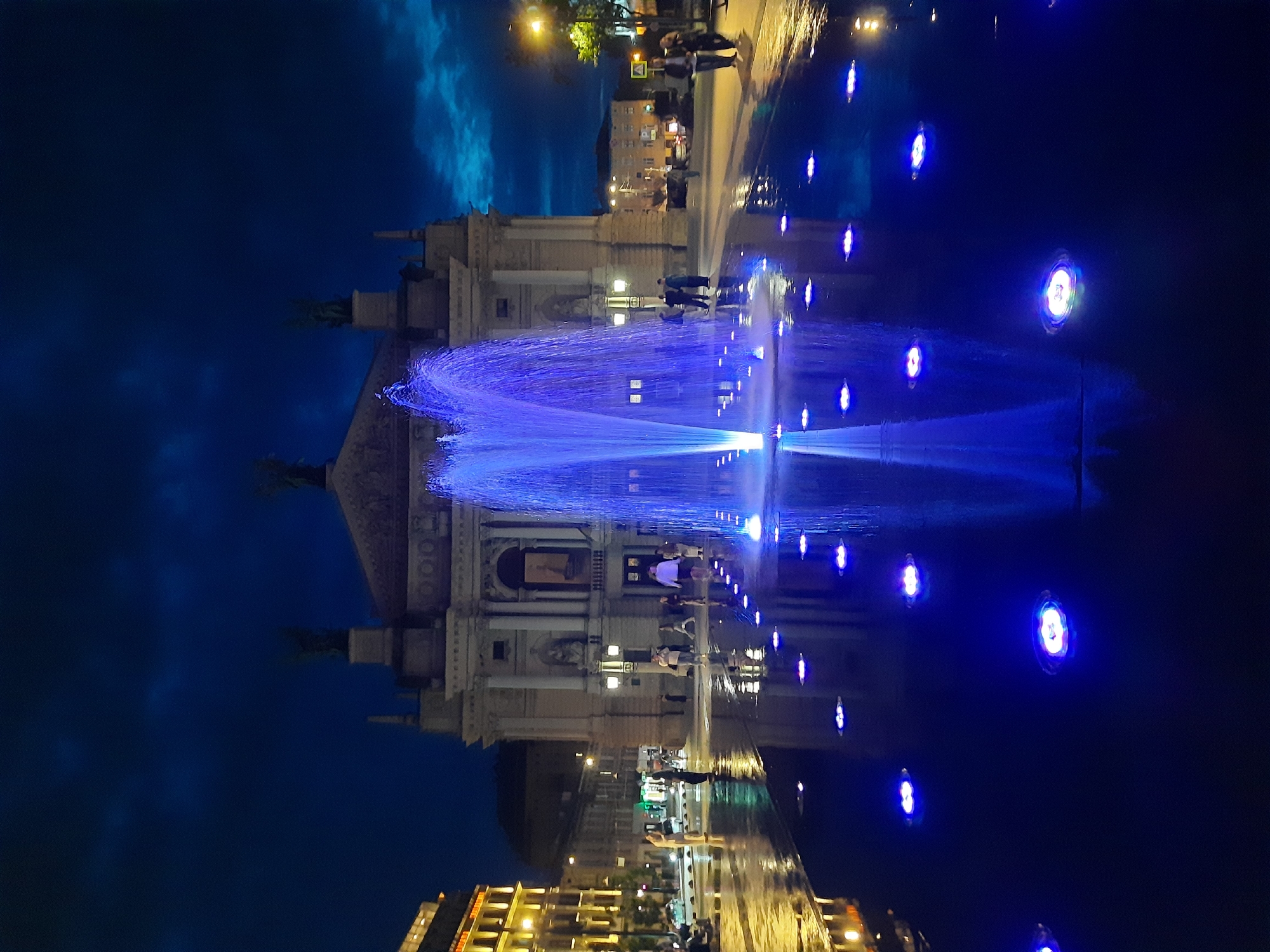 The image depicts a night scene in Lviv, Ukraine, specifically at the Rynok Square. In the foreground, a large, illuminated fountain is the central focus, with water jets reaching upwards and illuminated by bright blue lights. The fountain is situated on a wet, reflective surface, likely a water feature or a recently rained-on plaza, dotted with further blue lights submerged in the water.
In the midground, several people are visible, appearing to be tourists or locals enjoying the evening. They are standing and walking around the fountain and the plaza. Behind the fountain and the plaza stands a grand, classical-style building, likely an opera house or theater, with ornate architectural details and illuminated windows. The building's facade is lit, highlighting its structure and statues.
To the left of the building, a multi-story building with illuminated windows is visible, suggesting it is a commercial or residential area. The sky above is dark blue with some hints of clouds, indicating it is nighttime. Streetlights and other ambient lighting contribute to the overall illumination of the scene, creating a vibrant and atmospheric urban setting. The reflection of the fountain and the surrounding lights on the wet surface adds depth and visual interest to the composition.