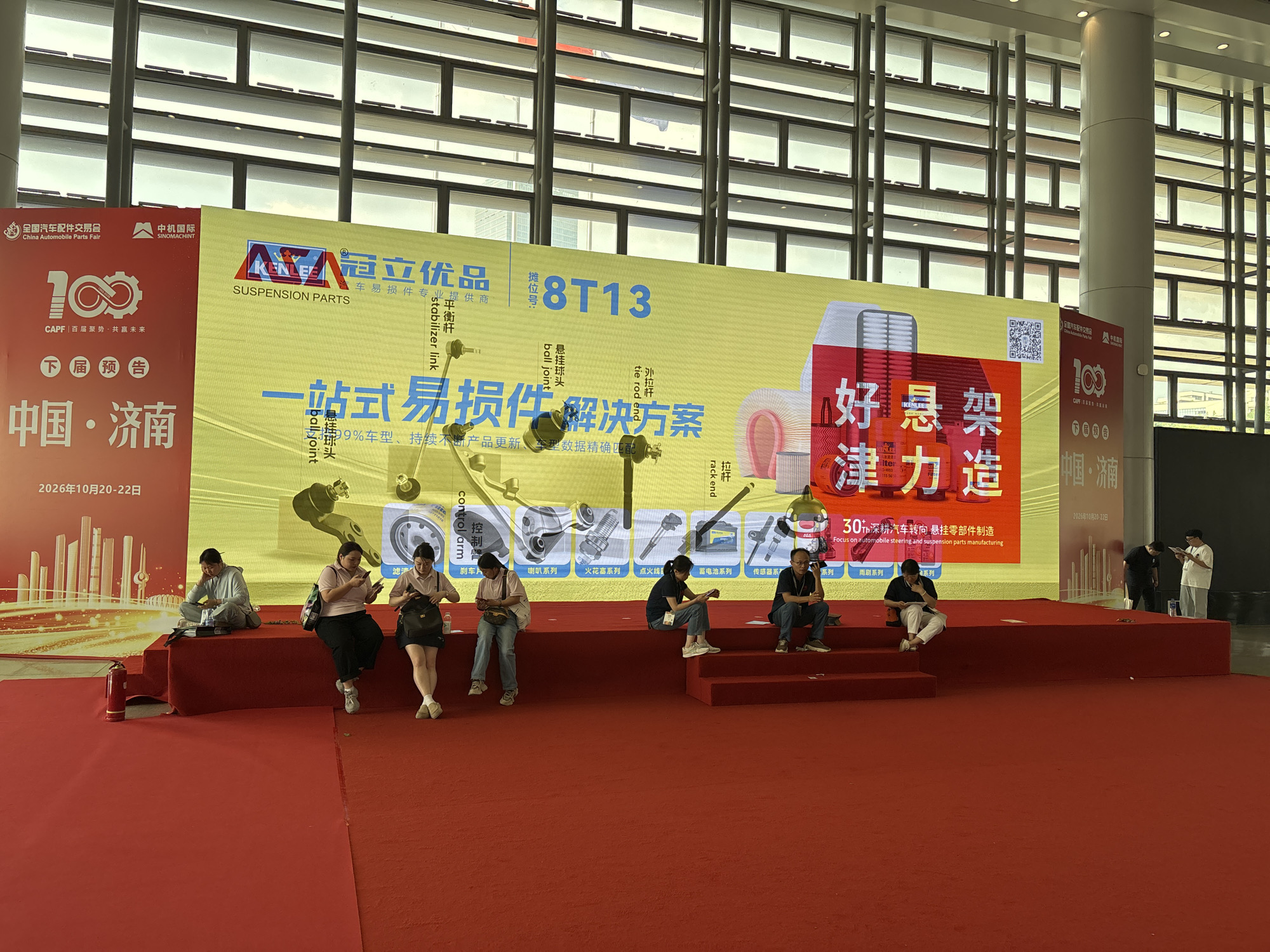 The image shows an indoor scene at an automobile parts fair in Liangjiang, China. Several people are seated on a red-carpeted platform in front of a large display screen with Chinese characters and images of car parts. The display promotes "Kenlee Suspension Parts" and mentions "30+ years focus on automobile steering and suspension parts manufacturing". The banner has the Chinese phrases "一站式易损件解决方案" (One-stop easy-to-damage parts solution) and "好悬架,津力造" (Good suspension, Jinli made). There is also a "100" logo associated with CAPF (China Automobile Parts Fair), marking a centenary. The signage indicates a future event to be held in China, specifically in the city of Jinan, in 2026, between October 20th and 22nd. The people on the platform are sitting casually, some using their phones. Behind the display is a wall with many windows. The environment suggests a convention center or large exhibition hall.