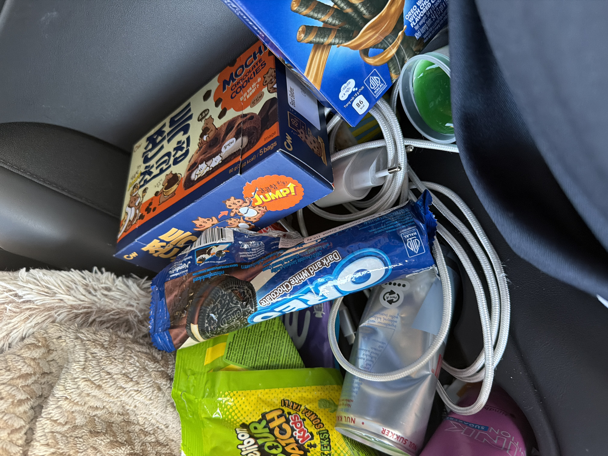 The image depicts a close-up, overhead view of various food items and electronic accessories cluttering a dark, textured surface, likely the seat of a vehicle. In the center, a blue box with Korean text and images of cookies is visible. To its left, a green bag of "Sour Patch Kids" candy is partially obscured. A blue package of "Oreo Dark and White Chocolate" cookies lies prominently in the foreground. Nearby, a white charging cable is coiled. To the right, a blue box of chocolate wafer rolls with a depiction of the rolls dipped in chocolate is present. A small, clear plastic container filled with green liquid is also visible. The background is dominated by a fuzzy, beige textile, possibly a blanket or car seat cover, and the dark, textured surface of the vehicle's interior. The scene appears to be inside a car, not in any specific location in Gothenburg, Sweden.