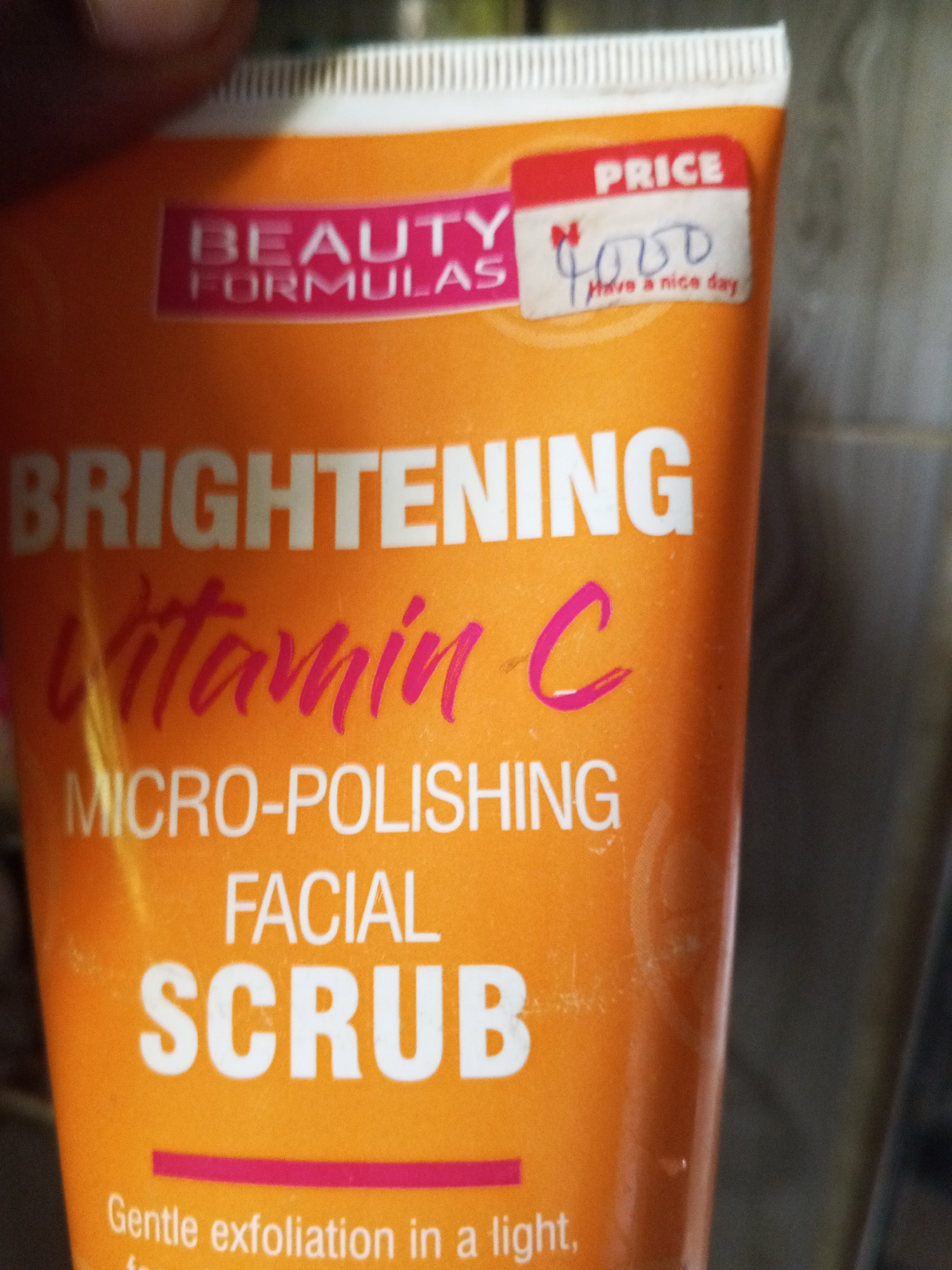The image shows a close-up of an orange tube of "BEAUTY FORMULAS" brightening Vitamin C micro-polishing facial scrub. A red price sticker is affixed to the tube, with "PRICE" written at the top and "N 4,000" handwritten below, followed by "Have a nice day." The product text is clear and in large font, with "BRIGHTENING VITAMIN C" being most prominent, followed by "MICRO-POLISHING FACIAL SCRUB." The bottom of the tube has a pink horizontal line and the beginning of text that reads "Gentle exfoliation in a light". The background is out of focus and appears to be a wall or surface. The image was taken in an indoor environment, likely a retail setting or a home. There are no people or specific landmarks that would indicate a location within a city.