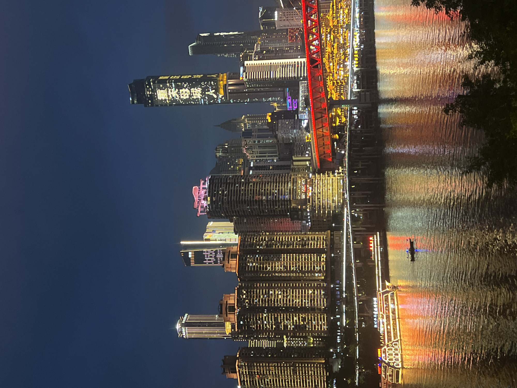 This is a nighttime photograph of a city skyline reflected in a body of water. The image is oriented vertically, with the skyline appearing to be upside down. The city features numerous tall buildings, many of which are illuminated with colorful lights. A prominent red bridge structure is visible, spanning across the skyline. The water is rippling, creating distorted reflections of the city lights. There is a small boat on the water, with a single person visible on it. The sky is dark blue, indicating it is nighttime. The overall mood of the image is serene and visually striking due to the interplay of light and reflection. The text "江北城街道" is visible on the side of a building.