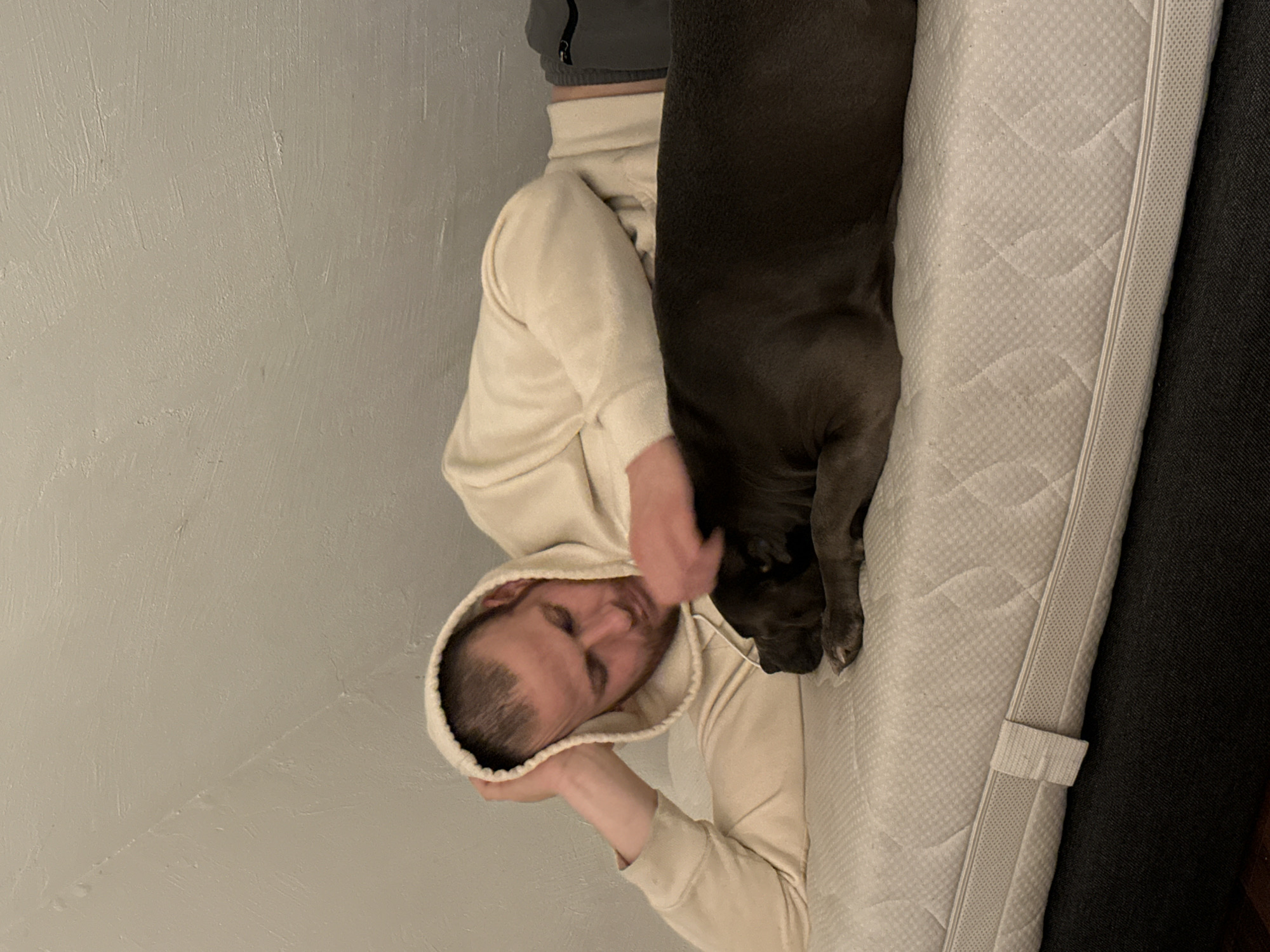 An adult male and a dog are resting on a mattress in an indoor setting in Heinebach, Germany. The male, wearing a light beige hooded sweatshirt and dark grey sweatpants, lies on his stomach with his head supported by his left hand. His right hand is placed on the back of a dark grey dog, which is also lying on its stomach to his right. Both subjects are positioned on a white quilted mattress with a visible side zipper and a fabric handle. A dark grey bed frame is beneath the mattress, and a light-colored, textured wall forms the background.