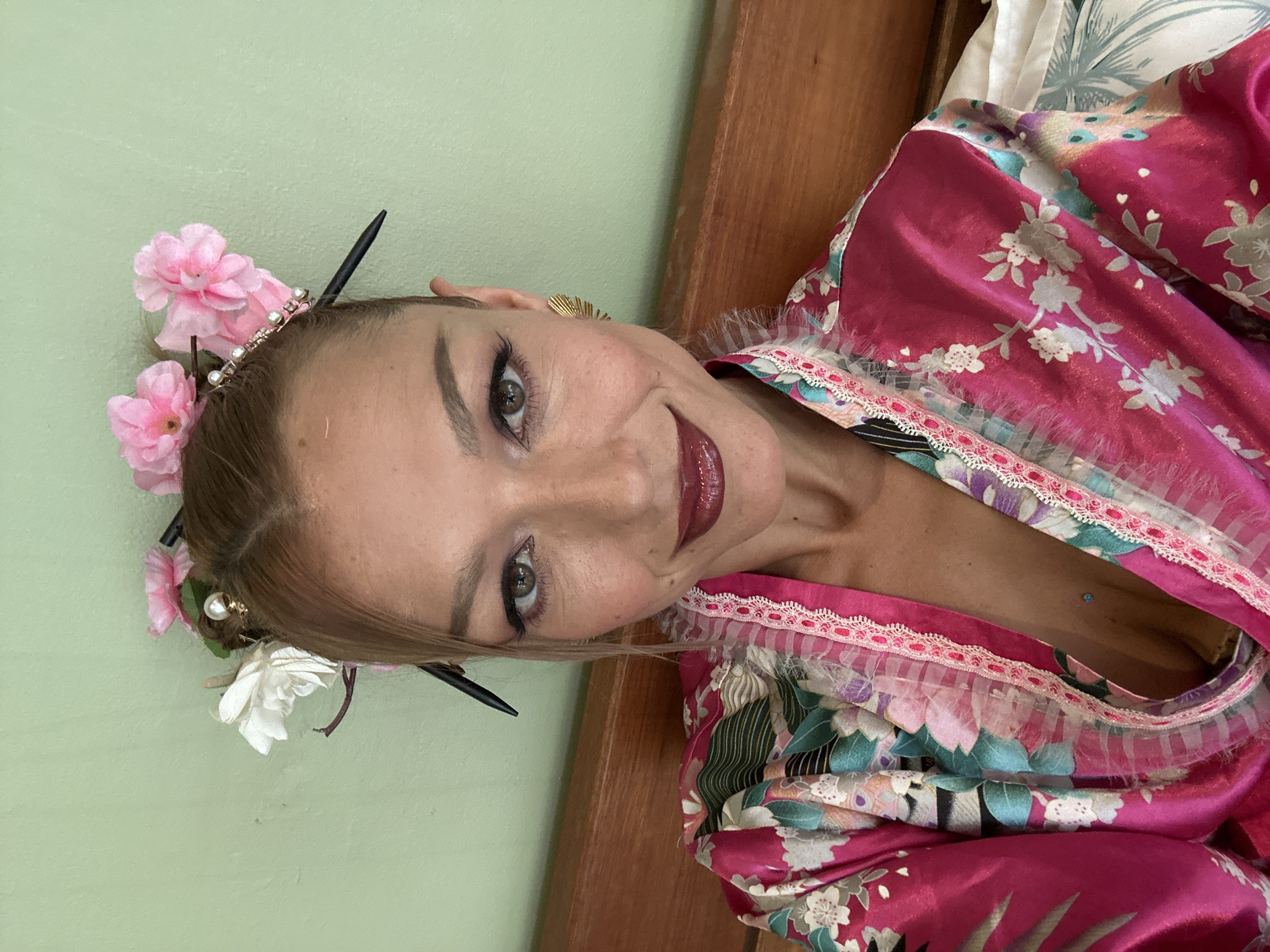 A woman is pictured in a close-up shot, wearing a vibrant pink kimono with a floral pattern and a lace trim. She has on dramatic winged eyeliner and dark, glossy lipstick, with a subtle piercing visible at the center of her chest. Her hair is styled with two buns, adorned with pink and white artificial flowers and two dark hair sticks. She is adorned with a golden, fan-shaped earring on her right ear. The background consists of a pale green wall and a wooden headboard, suggesting an indoor setting likely within a hotel room or residence in Ko Samui District, Thailand. The overall mood is one of self-expression and playful adornment.