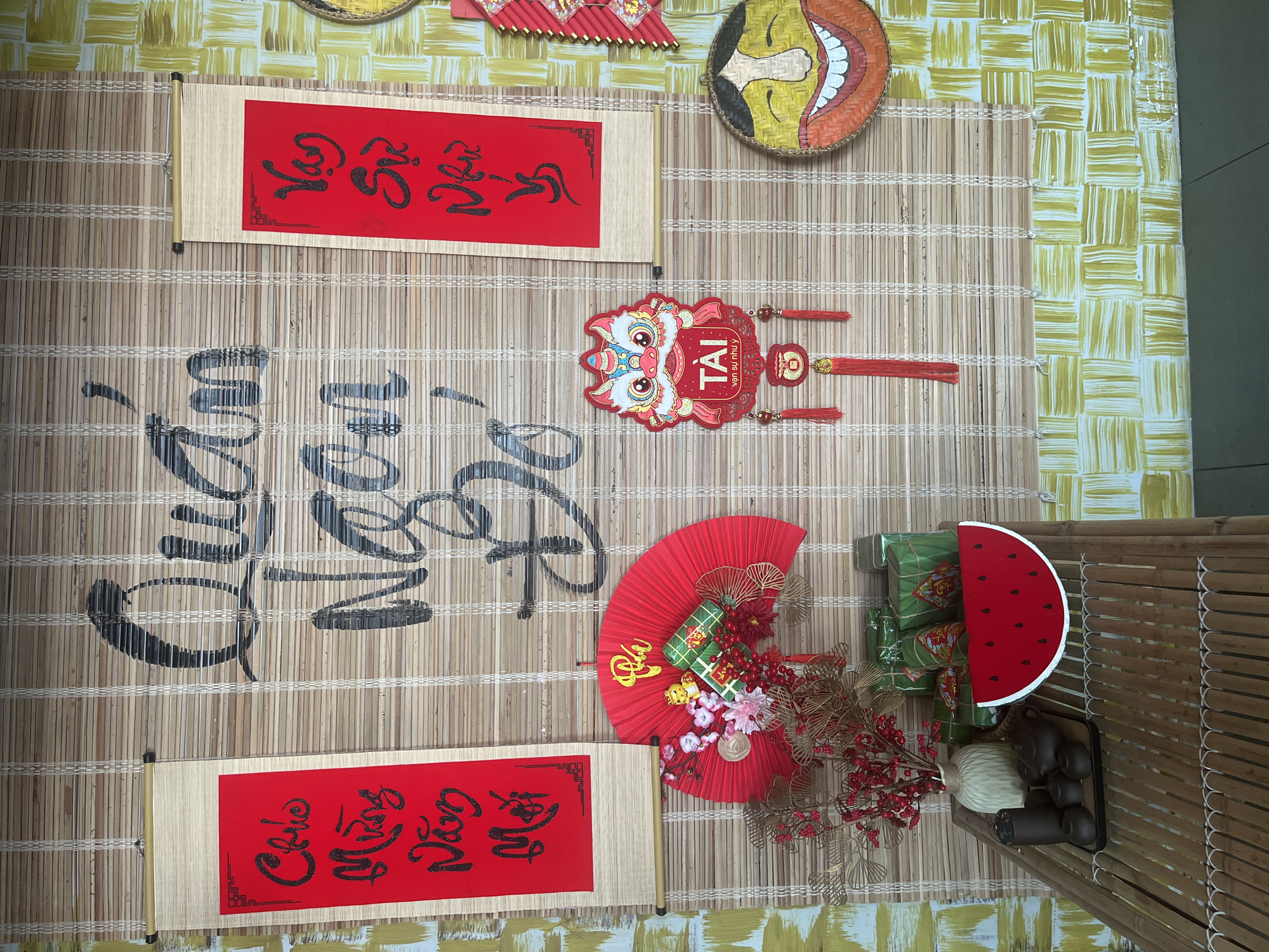 The image shows a decorative display, likely for the Vietnamese Lunar New Year (Tet). The scene is set on a bamboo mat with a yellow-and-white checkered background. The display includes:
1. Two red scrolls with black calligraphy, which are traditional Tet greetings.
2. A decorative lion head ornament with the word "Tài" (meaning wealth or prosperity).
3. A red fan decorated with Tet-themed items such as bánh chưng (sticky rice cakes), flowers, and gold decorations.
4. Bánh chưng wrapped in green leaves.
5. A watermelon slice prop.
6. A tea set placed on a small bamboo table.
The objects and arrangement suggest a festive occasion or display, likely set up in preparation for or during the Tet holiday. The scene's location and time of year are indicated by the Lunar New Year decorations. The specific city and country are Unknown.
