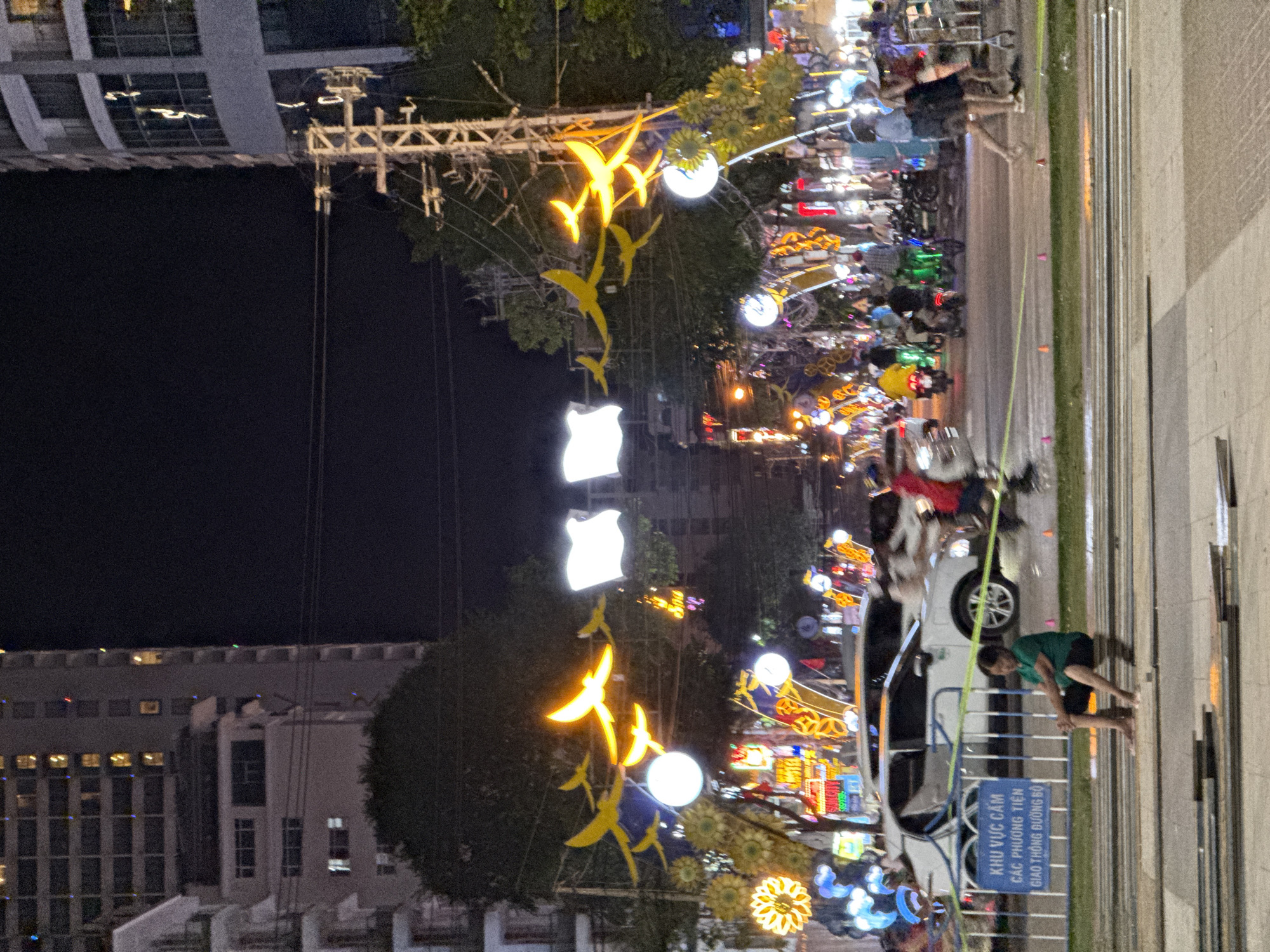 The image depicts a street scene at night in Nha Trang, Vietnam, likely during a festive period, evidenced by the abundant decorative lights. The foreground features a white car partially obscured by a metal barrier and a sign with Vietnamese text, indicating a restricted area for pedestrian traffic. A person wearing a green shirt and dark shorts sits on a low concrete structure in front of the barrier. Beyond the barrier, a street is bustling with activity, including several motorcycles and a few pedestrians. The street is lined with trees and tall buildings, with overhead power lines and elaborate light decorations, including illuminated birds and floral motifs, spanning across the road. The buildings are illuminated, suggesting they are hotels or apartments. The overall atmosphere is lively, with a mix of traffic, pedestrians, and festive decorations under the night sky.