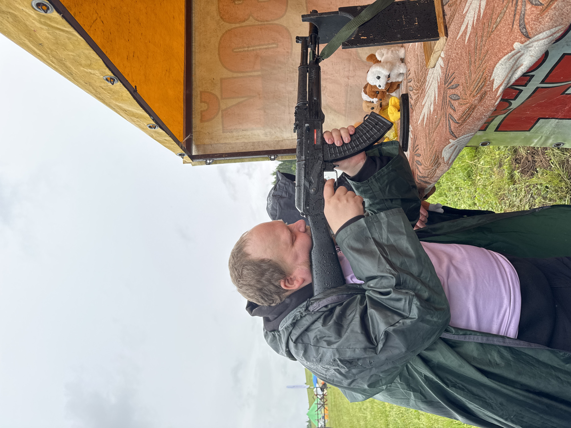 A man is depicted aiming a black rifle, resembling an airsoft or pellet AK-47, in an outdoor setting at Большая Кивара, Russia. He has short light brown hair and a beard, and is wearing a dark green hooded raincoat over a black hoodie and a light purple shirt. His head is pressed against the rifle's stock, with his eyes appearing closed or squinted in concentration. Water droplets are visible on his raincoat and the rifle, indicating damp or rainy weather.
The activity appears to be a shooting game or target practice, possibly at a carnival or festival. The man is positioned under a yellow and orange canopy or tent, which has partially visible red text that includes the letters "BON". To the right, another patterned banner, reddish-brown in color, displays partial red letters like "LA" or "TA". On a shelf behind the rifle, several small, light-colored stuffed animals and some yellow objects are arranged, likely prizes for the game. The ground is grassy, and in the distance, under an overcast sky, other colorful tents or structures are faintly visible. The scene is set during daytime, characterized by a bright but heavily clouded sky.