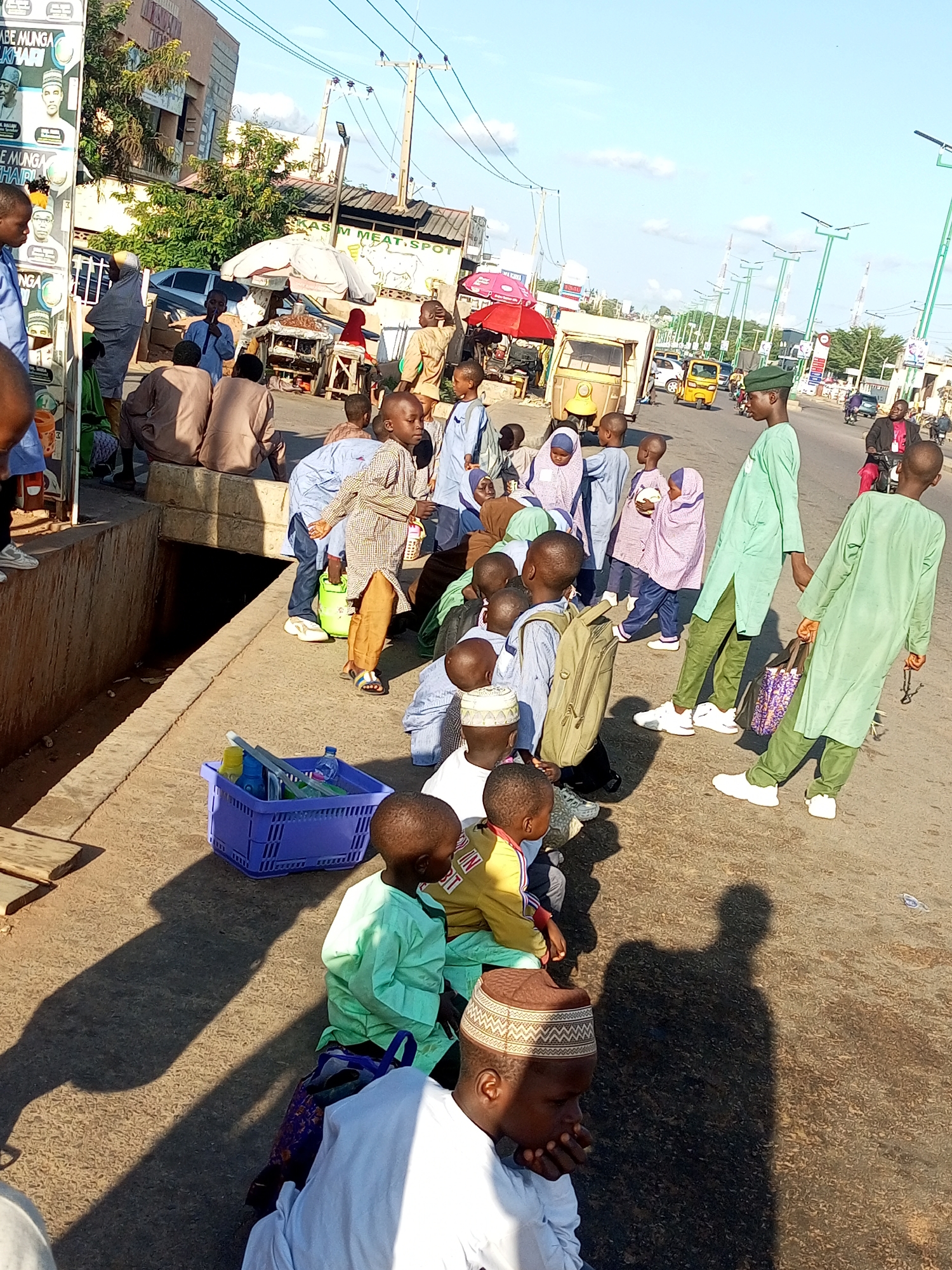 The image captures a street scene in Gombe, Nigeria, with a group of children, mostly boys, gathered on a concrete walkway and the adjacent road. The children are dressed in traditional Islamic attire, including caps and long robes. Some appear to be wearing school uniforms. They are in various poses, with some standing, some sitting, and one boy in the foreground looking down and resting his chin on his hand. A blue plastic basket filled with bottles and other items sits on the walkway. In the background, there are market stalls, buildings, vehicles including a yellow tricycle (Keke Napep), motorcycles, and an overcast sky. The overall impression is of a busy urban environment, possibly near a school or a place of worship.