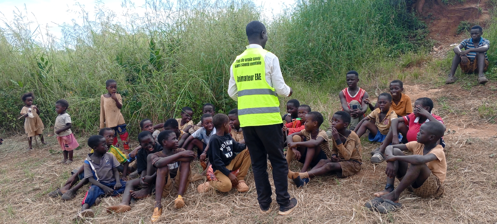 The image depicts a group of approximately 23 children and one adult in a grassy area in Ngalula, Democratic Republic of the Congo. The adult male is standing, facing the children, and wearing a white shirt and a neon yellow safety vest with text in French that reads "Pas de vraie santé sans santé mentale Animateur EAE". The children are mostly seated on the ground, looking at the adult. A few children stand on the left side of the frame. The vegetation consists of tall grass and shrubbery. A dirt embankment is visible in the background on the right. The environment appears rural, and the sky is overcast.