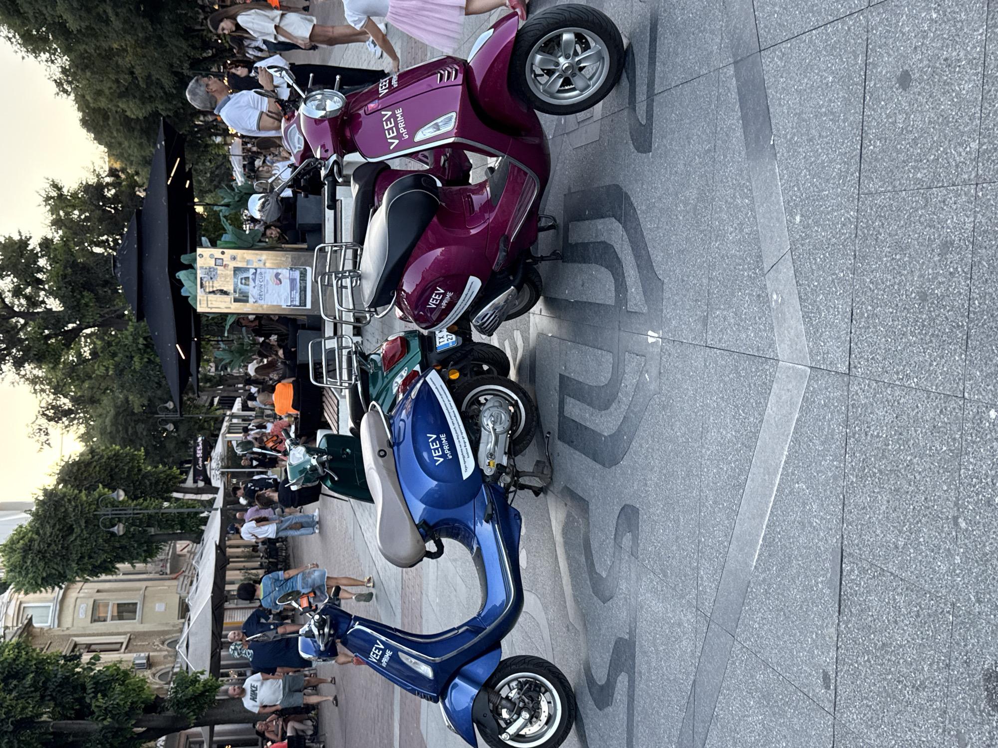 In this outdoor, urban scene, two vibrant scooters, one royal blue and the other a deep magenta, are parked prominently on a grey, patterned pavement. The blue scooter is on the left, while the magenta scooter is to its right, slightly behind. Both scooters feature the "VEEV in PRIME" logo. In the background, a bustling pedestrian area unfolds with people walking, seated at outdoor tables, and a variety of trees and buildings lining the street. The lighting suggests it is either early morning or late afternoon.