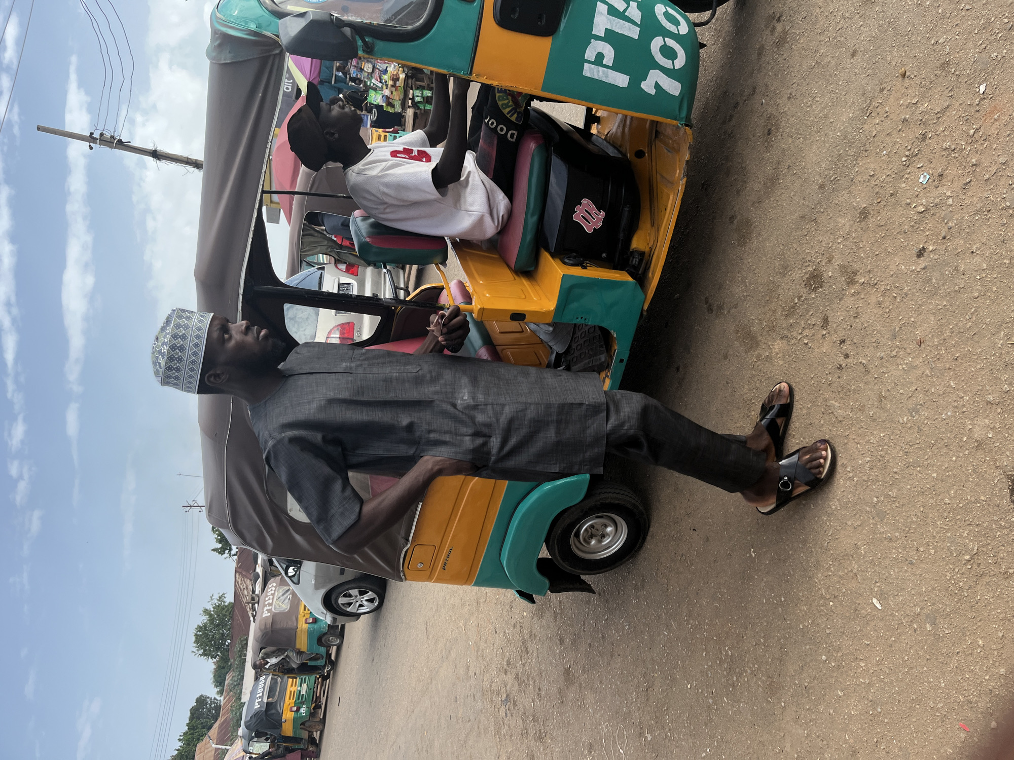 In Gura Top, Nigeria, a man wearing a dark grey traditional outfit and a patterned cap stands beside a yellow and teal auto-rickshaw. He has his left hand in his pocket and his right hand resting on the vehicle's roof. The auto-rickshaw has the partial text "PTA" and "700" on its side. Another man, wearing a white t-shirt and a dark baseball cap, is seated in the driver's position of the auto-rickshaw. Behind this auto-rickshaw, a silver car is visible, and further in the background, other auto-rickshaws are lined up on a dirt road. The scene is set under a partly cloudy sky, with buildings and power lines visible in the distance.