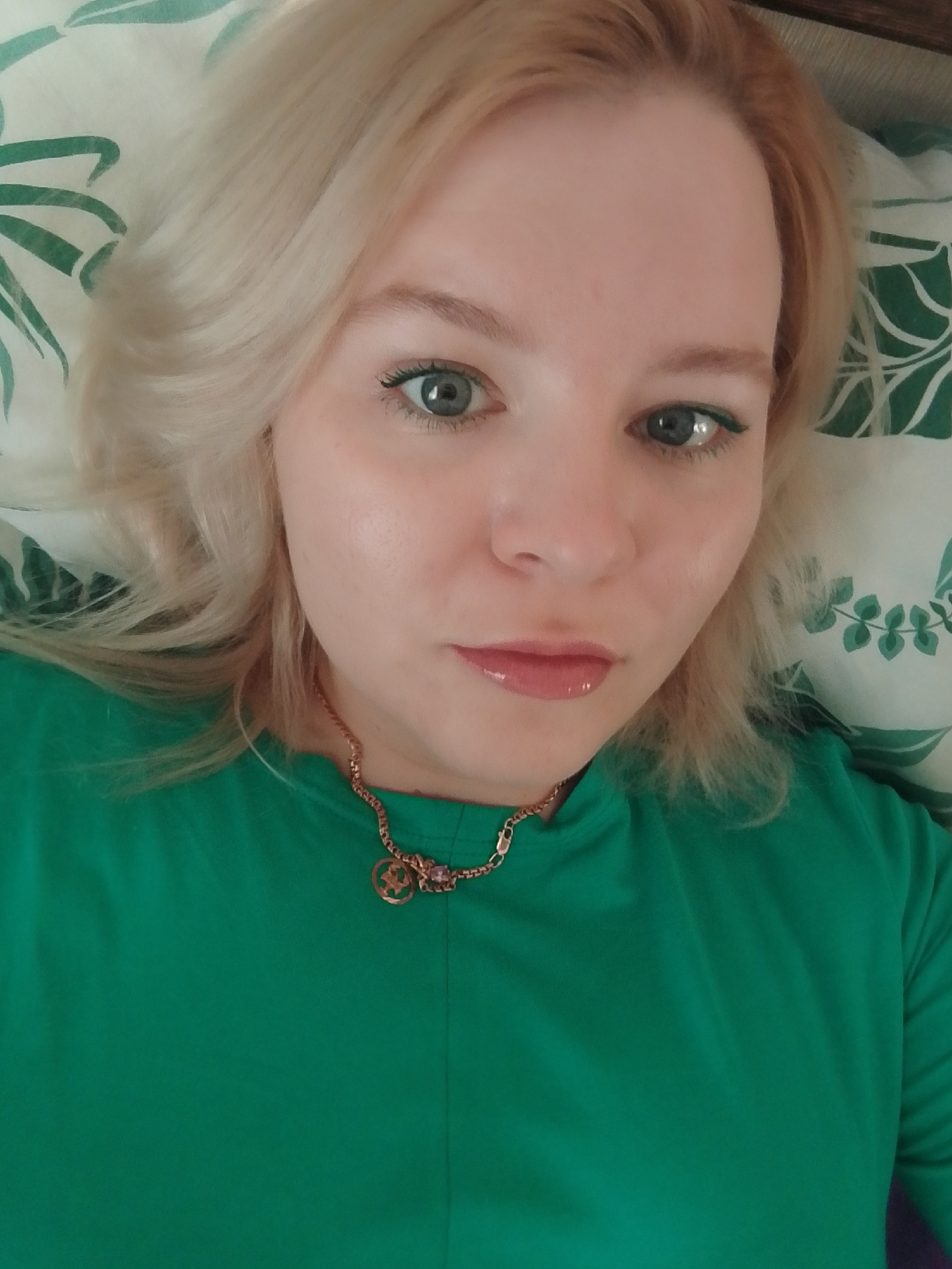 A close-up selfie depicts a fair-skinned, blond-haired woman with light eyes adorned with green eyeliner. She is wearing a bright green top and a gold necklace with a round pendant. The background shows a pillow with a green and white leaf pattern. There is no information present in the image to indicate a location in the city of Izhevsk, Russia.