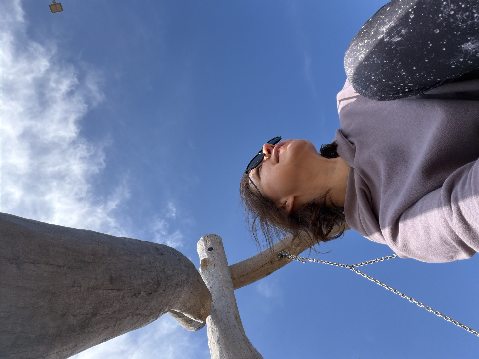 An extreme low-angle, upward-looking shot captures a person and large wooden structures against a bright blue sky with scattered white clouds. The person, likely female, is seen from the chest up, with dark hair, wearing a light purple/grey hoodie, and sunglasses perched on her nose. Her gaze is directed upwards, towards the sky. A dark, speckled object, possibly a bag or part of an accessory, rests on her upper arm.
To the left, a thick, horizontal wooden log-like structure extends across the frame. Below it, a vertical wooden post with a crossbar is visible, from which a metal chain hangs, extending towards the bottom right. These wooden elements, along with the chain, suggest the setting is a playground or an outdoor recreational area, possibly involving a swing or similar equipment. High in the top left corner, a small, square, light-colored object, possibly a distant light fixture, is visible against the sky.
The scene is set outdoors during bright daylight, indicated by the clear blue sky and strong illumination, suggesting good weather. The location context is Chaikovsky, Russia. No visible text is present in the image.