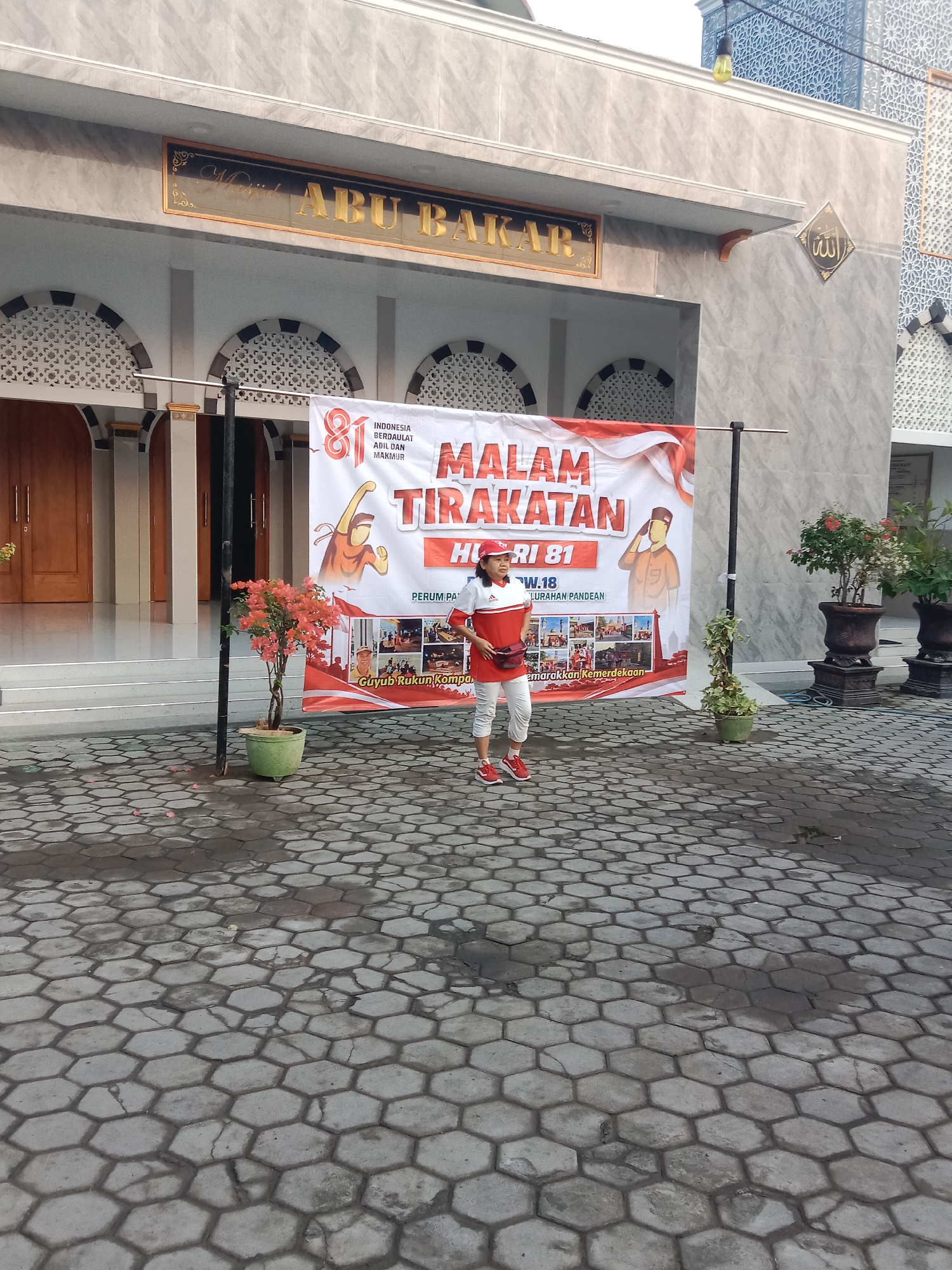 A woman in athletic attire, including a red cap and white and red shirt and pants, stands in front of a banner in the city of Madiun, Indonesia. The banner features text in Indonesian, including "MALAM TIRAKATAN HUT RI 81" and "INDONESIA BERDAULAT ADIL DAN MAKMUR". The woman appears to be participating in an event, possibly a celebration related to Indonesian Independence Day. Behind her is a building with Islamic architectural elements, identified by a sign as "MASJID ABU BAKAR". The ground is paved with hexagonal stones, and there are potted plants and large planters with flowers present. The lighting suggests it is daytime.