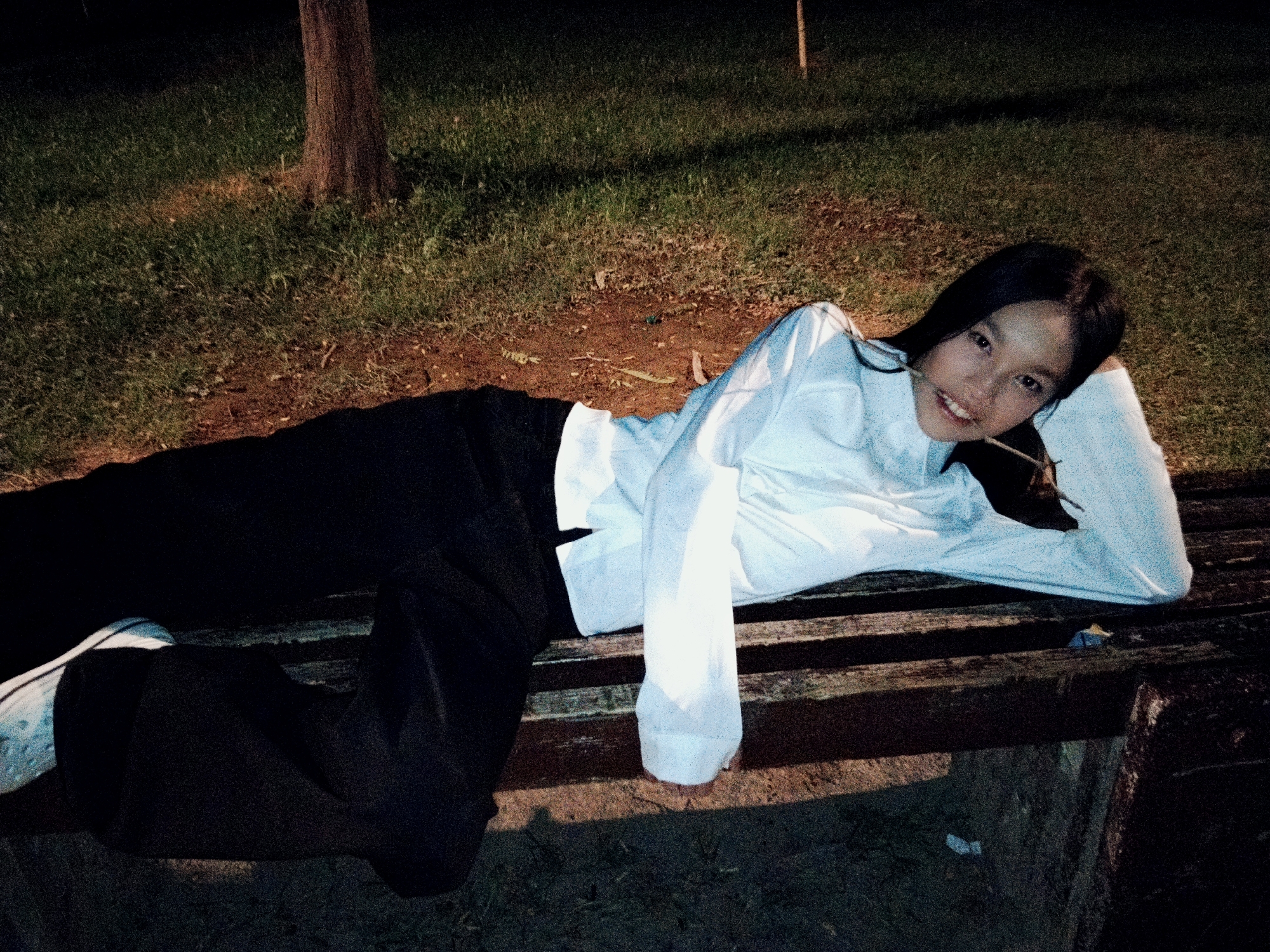 A young woman with long dark hair is lying on her side on a wooden park bench. She is wearing a white long-sleeved shirt and black pants, with white shoes visible on her left foot. Her right arm is bent, with her hand supporting her head, and she is holding a thin twig in her mouth. She is smiling towards the camera. The bench is situated on a patch of grass next to a dirt path, with a tree trunk visible in the background to the left. The overall lighting suggests it is either dusk or nighttime. The scene appears to be a park in Bishkek City, Kyrgyzstan.