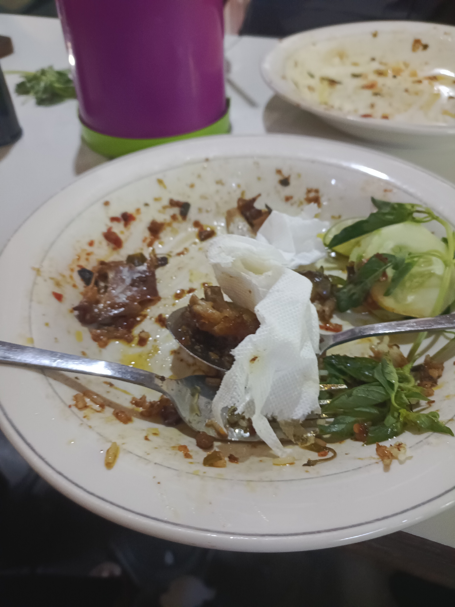 The image presents an indoor dining scene in Janti, Indonesia, depicting the aftermath of a meal. A central white ceramic plate, detailed with a thin grey line along its rim, occupies the majority of the lower frame. This plate contains substantial food residue, including dark brown sauce remnants, scattered reddish spice fragments, and visible oil stains. A silver fork lies on the right side of the plate, partially beneath a small cluster of fresh green basil leaves and several slices of light green cucumber. A silver spoon rests on the left, holding a piece of cooked meat or poultry. Two crumpled white paper napkins are situated near the center of the plate, one partially obscuring the spoon. In the upper right background, a second white ceramic plate, out of focus, shows sparse food remnants. To the upper left, a vibrant purple cup with a distinctive lime green base stands on a white table surface. A small cluster of fresh green leaves is visible next to the cup. The scene is illuminated by ambient indoor lighting.
