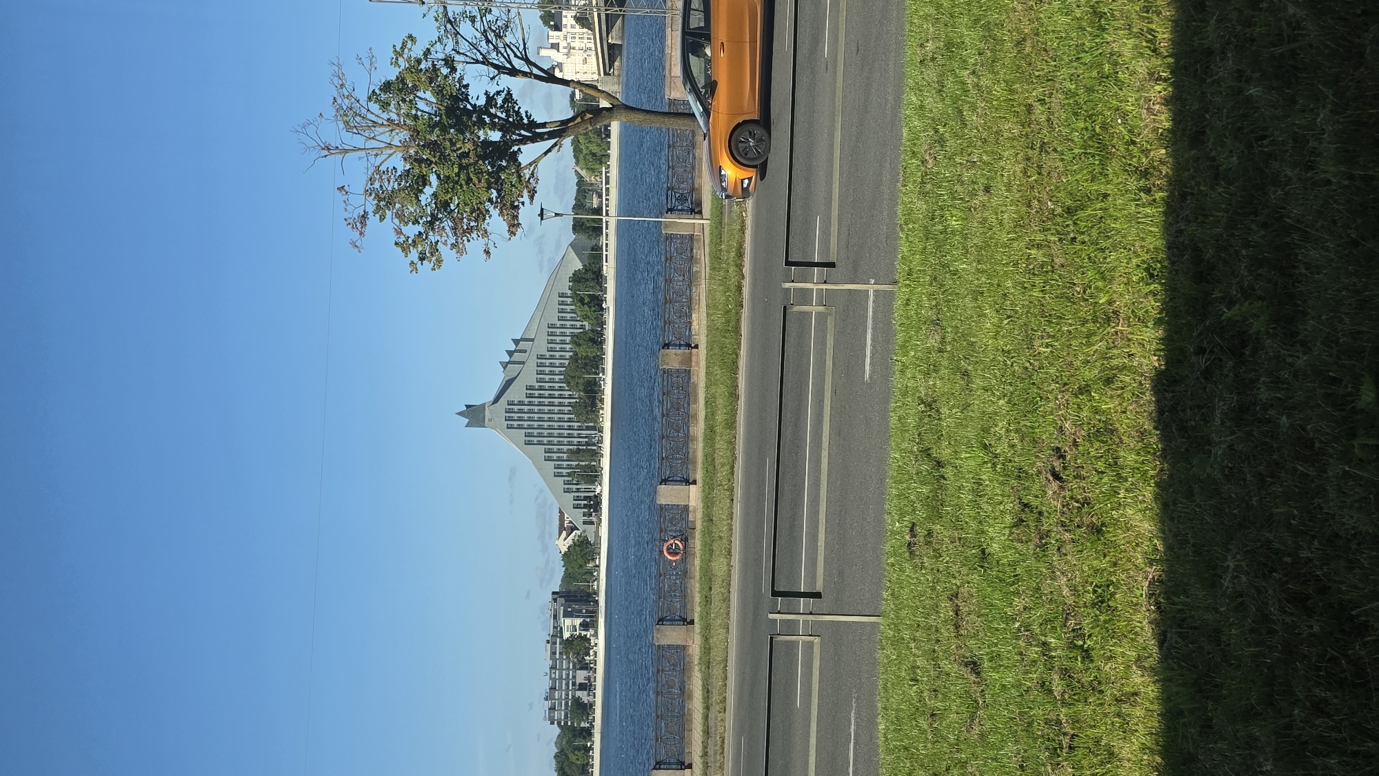 An orange car is driving on a road in front of a river in Riga, Latvia. The Latvian National Library is in the background. The sky is clear and blue. The car is the only vehicle visible on the road. The river is calm and reflects the blue sky. The library is a modern building with a distinctive angular roof. There is a tree on the right side of the image. There is a grassy area in the foreground.