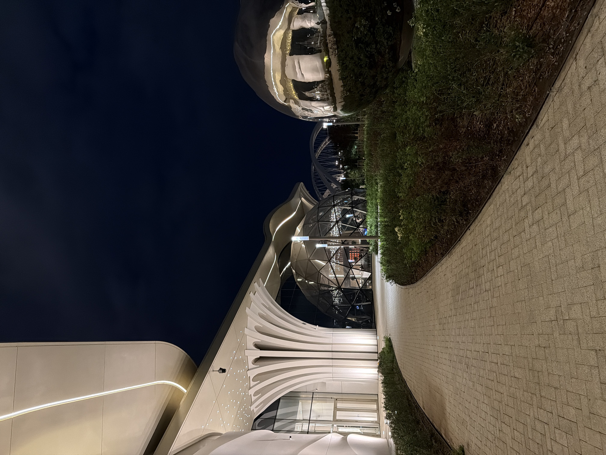 This is a nighttime, horizontal shot of a modern architectural complex in Astana, Kazakhstan. The scene features a sleek, white building with curved lines and integrated lighting, adjacent to a geodesic dome structure illuminated from within. To the left, a large, reflective spherical sculpture is partially visible, mirroring the surroundings. A paved walkway, laid with light-colored rectangular stones in a herringbone pattern, curves through the foreground, bordered by lush, manicured greenery. In the background, a modern bridge structure with intricate metalwork is illuminated against the dark night sky. The lighting suggests a cool, clear evening. The overall ambiance is one of contemporary design and sophisticated urban planning. There is no visible text in the image.