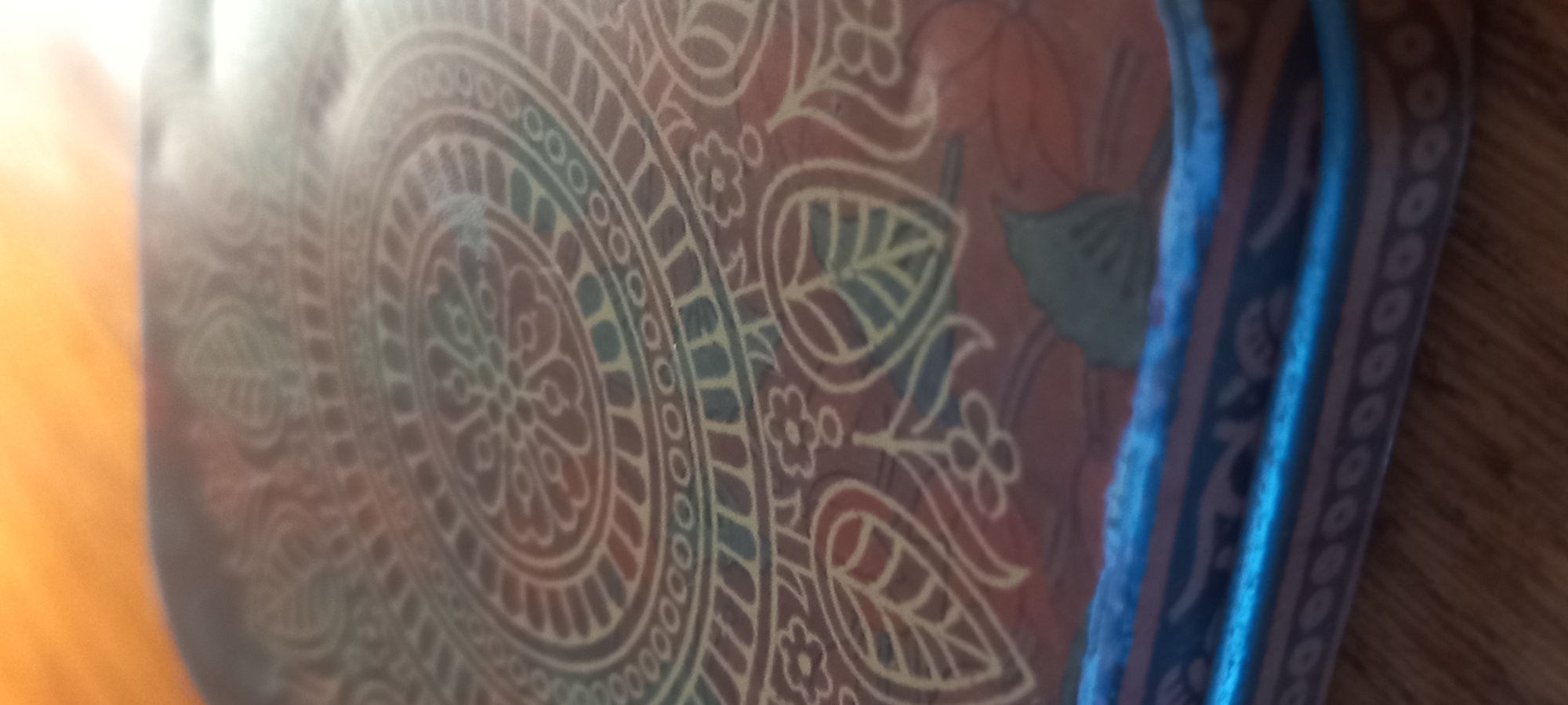 The image shows a close-up, angled view of a circular, decorative object, possibly a cushion or a lid, with an intricate mandala-like pattern. The pattern features concentric circles filled with floral and leaf motifs in shades of red, brown, blue, and yellow. The object has a blue and striped edge. The background is blurred, showing a wooden surface, likely a table or floor, and an orange-toned area that could be a wall or another surface. There are no people or any discernible city landmarks visible.