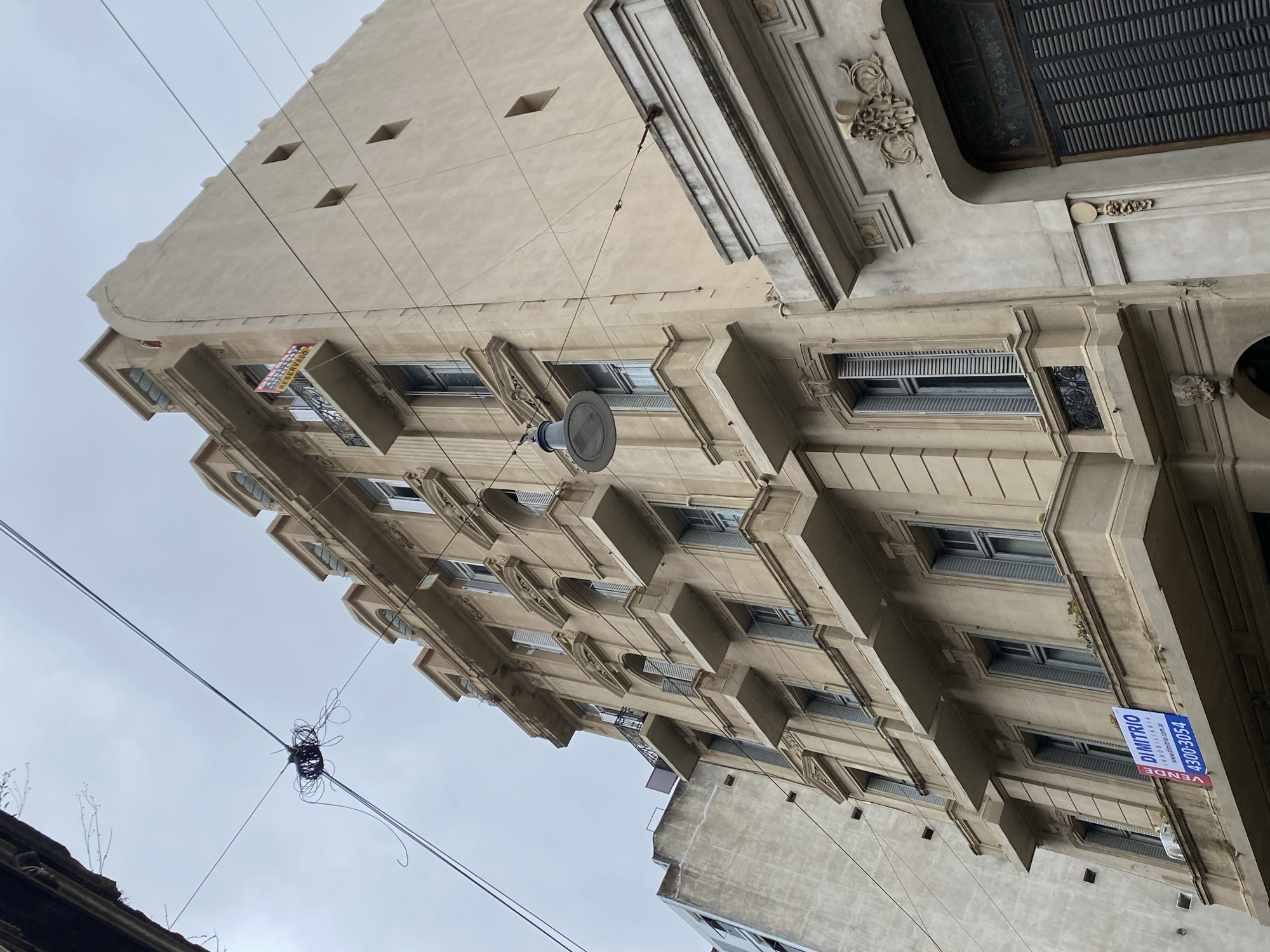 The image shows a low-angle view of a multi-story building in Buenos Aires, Argentina. The building features a beige facade with ornate architectural details, including protruding window frames, decorative moldings, and a top-level cornice. A blue and white "Vende" ("For Sale") sign is attached to one of the lower floors. Overhead wires are visible in the foreground, along with a street lamp and a tangled mess of cables. The sky is overcast, suggesting a cloudy day. The other building is a concrete building.