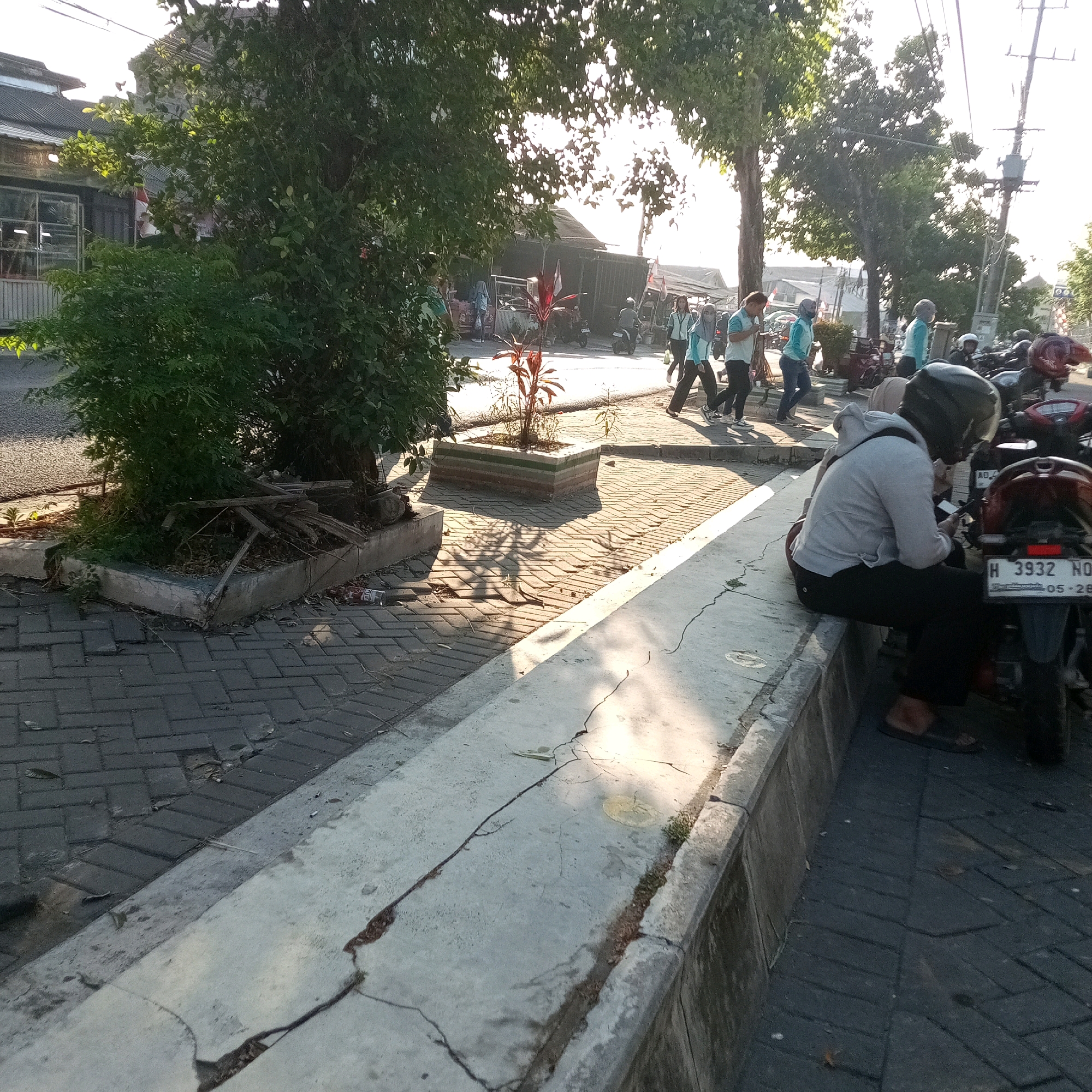 The image depicts a street scene in Butuh, Indonesia, likely during daylight hours given the strong sunlight and shadows. In the foreground, a person wearing a grey hoodie and black pants sits on a concrete curb, looking at a mobile phone. They are seated next to a dark-colored motorcycle with a visible license plate that reads "H 3932 NO" and "05 - 28". To the left of the seated person, the curb continues, covered in cracked concrete, with debris and a discarded plastic bottle. The ground in the foreground is paved with dark grey brickwork.
Further into the scene, a tree with dense green foliage casts long shadows across the brick pavement. Several other people, some wearing light blue shirts and dark pants or hijabs, are walking or standing in the background. They appear to be moving along a paved walkway or street. Various buildings and shops, some with metal roofs and signage, are visible behind the people. Multiple motorcycles are parked along the street and in front of the shops. The overall environment appears to be a commercial or public area within the city.