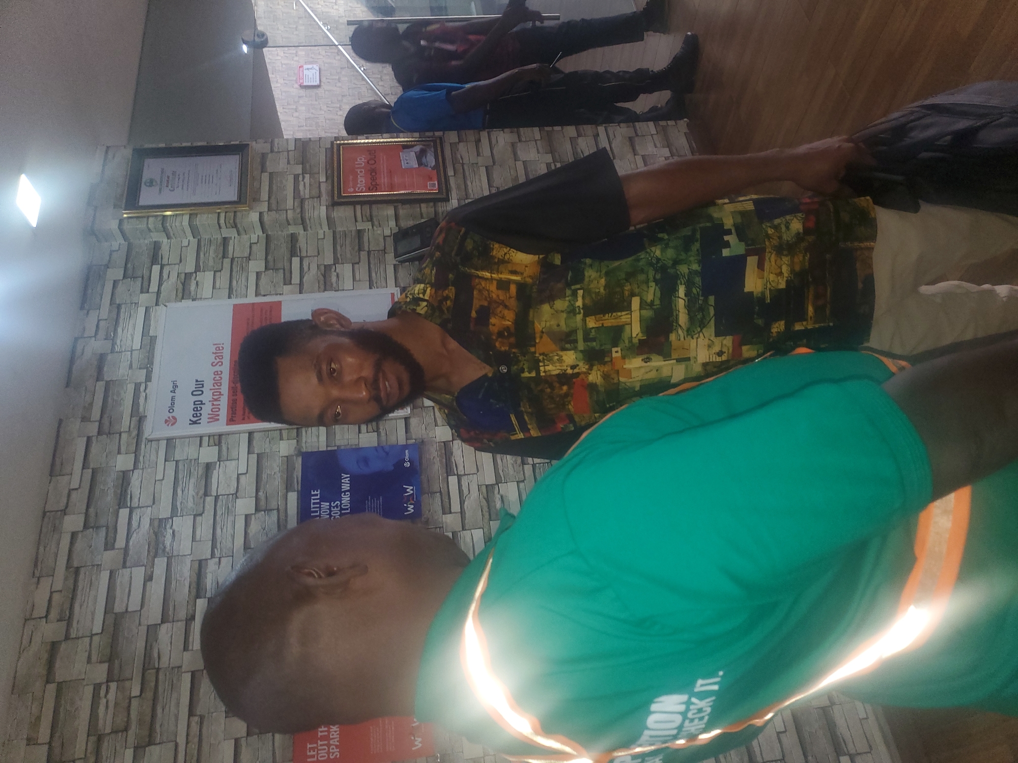 Two men are seen standing indoors, facing each other in conversation. The man on the left wears a bright green shirt with orange reflective stripes and appears to be a worker. The man on the right wears a colorful, patterned shirt and holds a black bag. The background features a textured, grey brick-like wall with framed posters and certificates hanging on it. The floor is made of polished wood. In the background, other individuals can be seen walking towards a staircase. The scene suggests an indoor public space, possibly an office or a common area within a building.