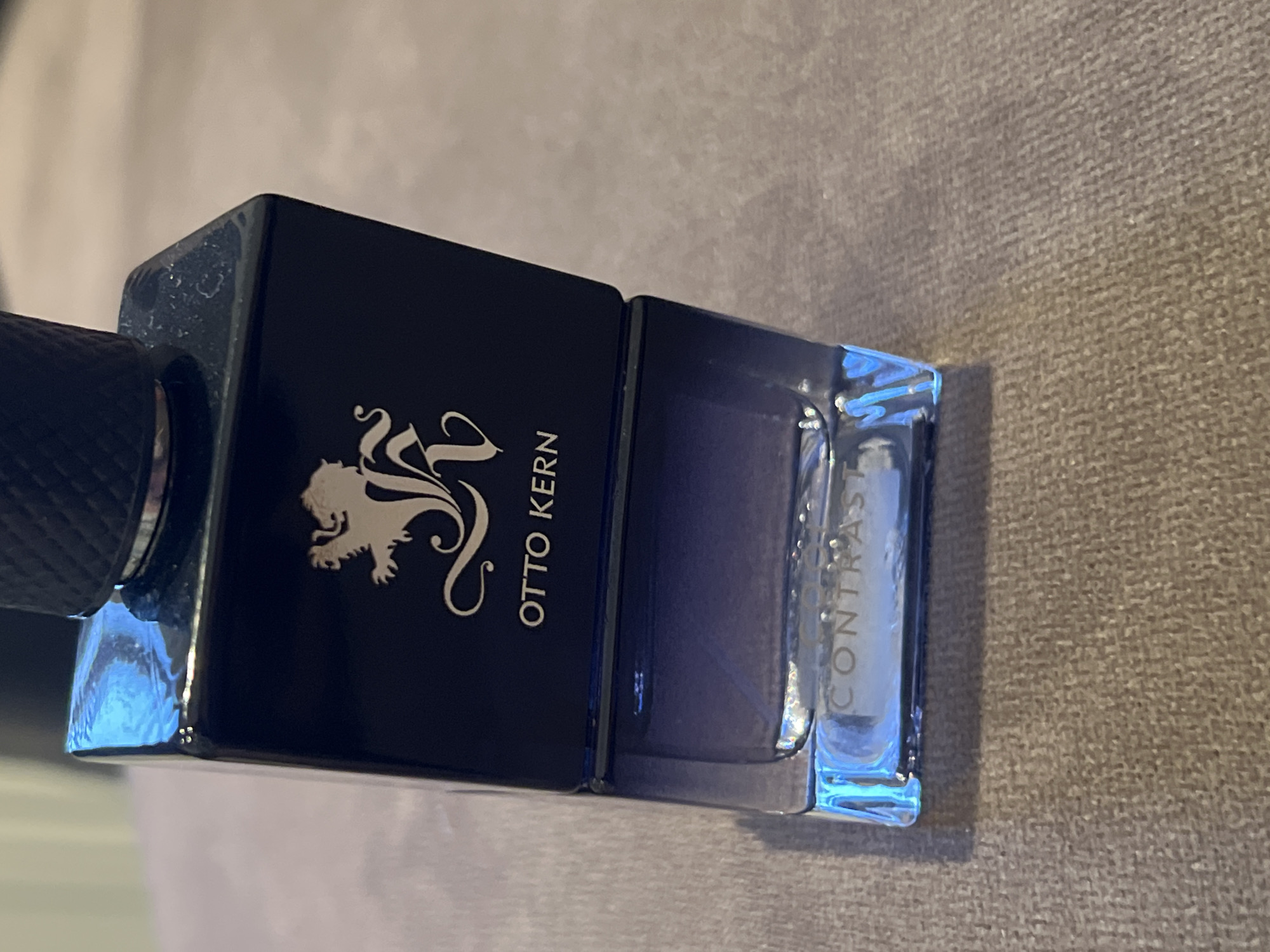 A black glass perfume bottle with a silver lion emblem and "OTTO KERN" text sits on a textured brown surface. The lower half of the bottle is clear glass, revealing a gradient of purple liquid. The words "CONTRAST" are visible etched into the bottom of the bottle. The image is taken from a slightly elevated angle, focusing on the bottle.