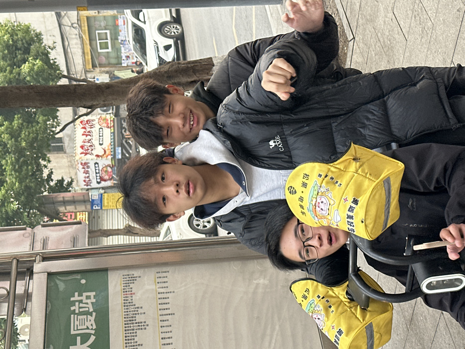 Three young East Asian males are positioned closely together in an urban street scene in Jiangjin, China, near the "Dasha Station" bus stop. In the immediate foreground, a male with glasses and a dark long-sleeved shirt is seated, operating an electric scooter or bicycle. His hands grip the handlebars, which are fitted with bright yellow, padded covers adorned with cartoon characters and Chinese text, including "东城美滋" and "雨洁・电・电". He looks towards the camera with an open-mouthed expression. Directly behind him, a male with short, curly dark hair, wearing a grey hooded sweatshirt under a black puffer jacket with a "CAMEL" logo, looks straight into the camera with a neutral expression. His left arm is bent, and his hand is partially visible in a gesturing posture. To the right and slightly behind this central figure, a third male with similar curly dark hair and a dark puffer jacket smiles widely, looking slightly to the right of the frame. The background features a street with a white sedan and a white SUV, commercial buildings displaying various Chinese signs including a red banner for "烤鱼火锅", and a deciduous tree. A bus stop sign with the characters "大厦站" is prominently visible on the left edge of the frame. The lighting indicates daylight conditions.