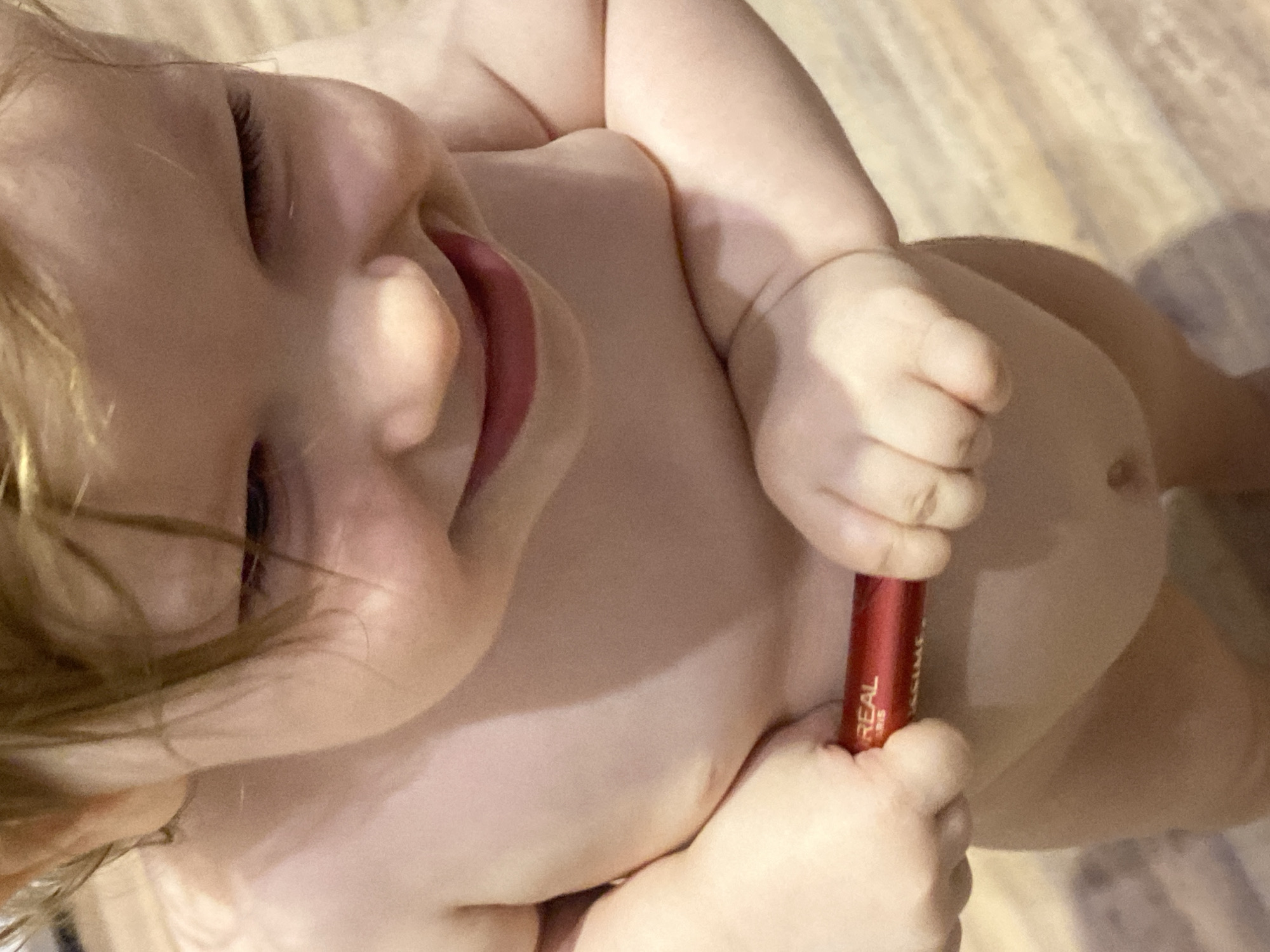 A joyful young child, appearing to be a toddler with light-colored hair and fair skin, is captured in a top-down, slightly angled shot. The child is smiling broadly, with rosy lips and visible eyelashes, suggesting a happy and playful mood. They appear to be lying down, possibly on a floor or bed, which is covered with a light-colored, textured surface resembling wood planks or light-toned laminate flooring.
The primary activity is the child holding a red cosmetic tube. Their small hands grasp the tube firmly against their bare torso. Visible text on the tube clearly identifies it as a L'Oréal product, with "REAL" and "L'ORÉAL PARIS" discernible, along with a partial view of "MAXIME". Given the context of a child playing, it's likely a tube of mascara or lipstick they are exploring.
The setting appears to be indoors, likely in a home in Krasnodar, Russia. The lighting is soft and even, suggesting either an indoor light source or indirect daylight. The child is mostly bare-chested, emphasizing their youth and playful interaction with the object. The overall impression is one of innocent curiosity and happiness.