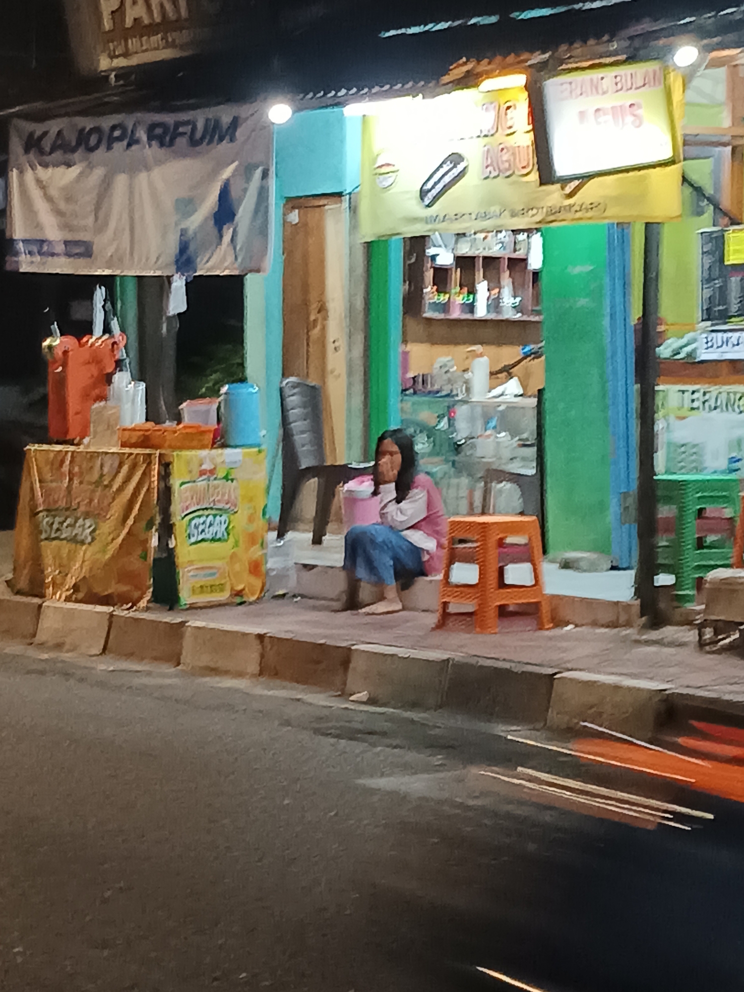 The image depicts a street-level view at night in the city of Yogyakarta, Indonesia, likely a commercial area. In the foreground, a blurry street with moving lights suggests passing vehicles. The midground features a sidewalk with a young woman sitting on an orange stool. She is wearing a pink jacket and blue jeans and appears to be covering her mouth with her hand, looking downwards. Beside her is a stall with two orange containers and a blue bucket. To the left of the stall, another, more draped stall is visible with the word "SEGAR" (fresh) printed on it. Behind the woman, a store facade is visible with shelves displaying various items. A banner overhead reads "KAJOPARFUM" and another sign displays "TERANG BULAN". The overall scene suggests a humble, possibly informal, commercial establishment operating at night.
