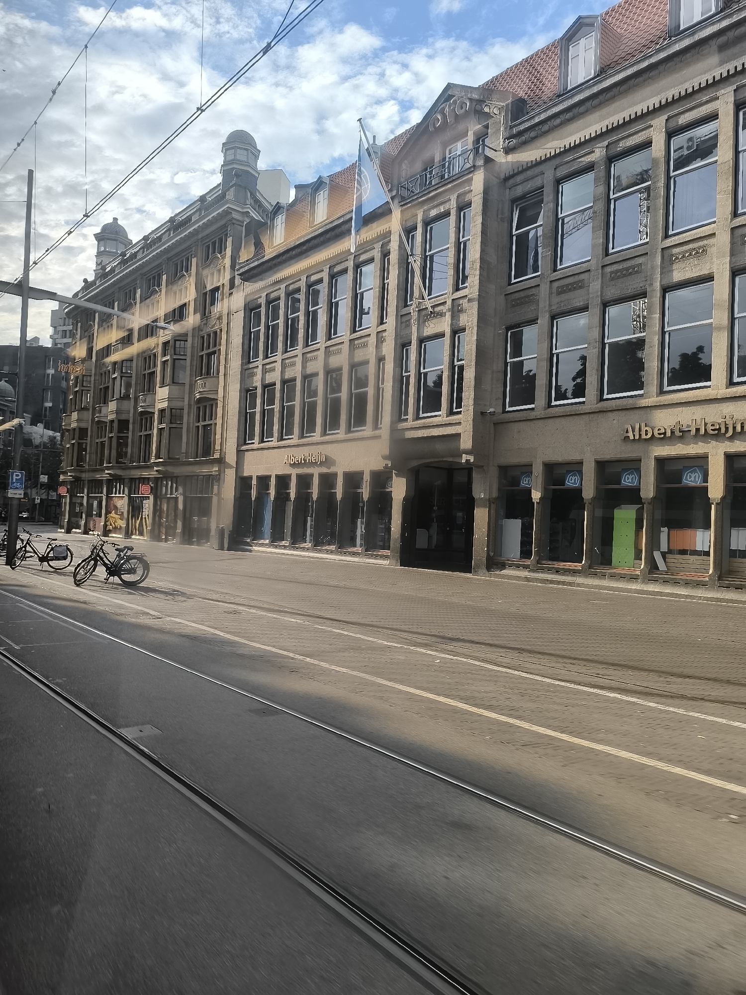 The image captures a street scene in The Hague, Netherlands. In the foreground, tram tracks run parallel to the street. The street itself is paved, with some sections showing cobblestones and others asphalt. Two bicycles are parked on the sidewalk on the left side of the image.
Dominating the background is a multi-story building with a stone facade. The building has numerous large windows, many of which appear to be reflective, showing the sky and surrounding structures. The ground floor of the building houses a supermarket with the "Albert Heijn" logo visible above the entrance. Several windows on the ground floor display interior views of the store, with some showing shelves and merchandise. The building's upper floors feature a more ornate architectural style with decorative elements and varied window designs. Above the building, a cloudy sky with patches of blue can be seen. Overhead power lines are also present, suggesting the presence of public transportation, likely trams or trolleybuses. The overall impression is of a typical urban street with shops and infrastructure.