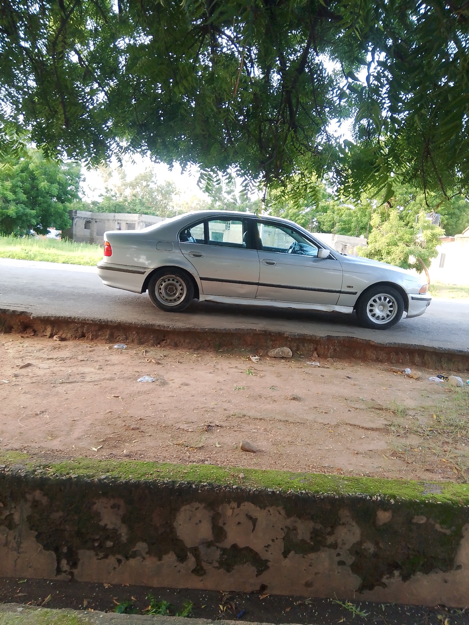 The image shows a silver sedan parked on the side of a road. The car is positioned to the right of the frame, facing left. It is a daytime scene, with the sun casting shadows and illuminating the foliage of a large tree that overhangs the car and the road. The tree's leaves are a vibrant green, suggesting it is a healthy, mature tree, possibly an acacia or neem tree common in Nigeria.
The car appears to be a BMW, likely from the late 1990s or early 2000s based on its body style. It is a four-door sedan with tinted windows. The wheels are silver alloy rims. The car is parked on an asphalt road that appears to be in fair condition, with some visible wear and tear.
In the foreground, there is a dirt embankment with some rocks and debris. The embankment is covered with some sparse green vegetation along its top edge. Further in the background, beyond the road, are more trees and some buildings. These buildings appear to be made of concrete and have a rustic, somewhat unfinished look, with visible windows and rooflines. One of the buildings has a water tank on its roof.
The overall setting suggests a somewhat rural or semi-urban environment in Jalingo, Nigeria. The presence of the large tree and the style of the buildings are consistent with this location. The lighting suggests it is daytime, likely late morning or early afternoon, given the angle of the shadows. There are no people visible in the image. The atmosphere is tranquil, with the focus being on the car and its surroundings. No text is visible in the image.